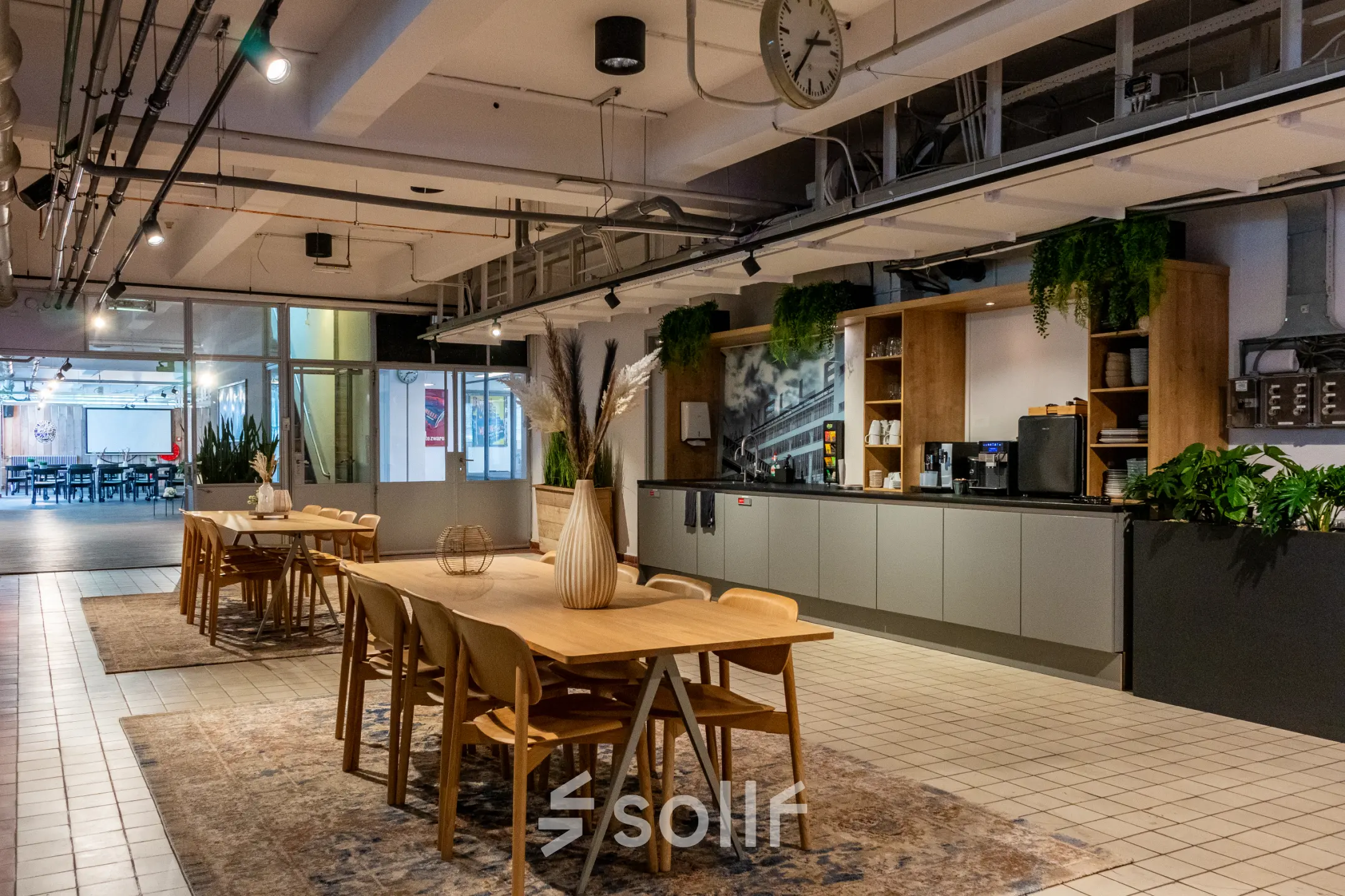 A modern office space at Van Nelleweg 1, Rotterdam Delfshaven, featuring wooden tables, chairs, potted plants, and a kitchenette, ideal for office space rental.