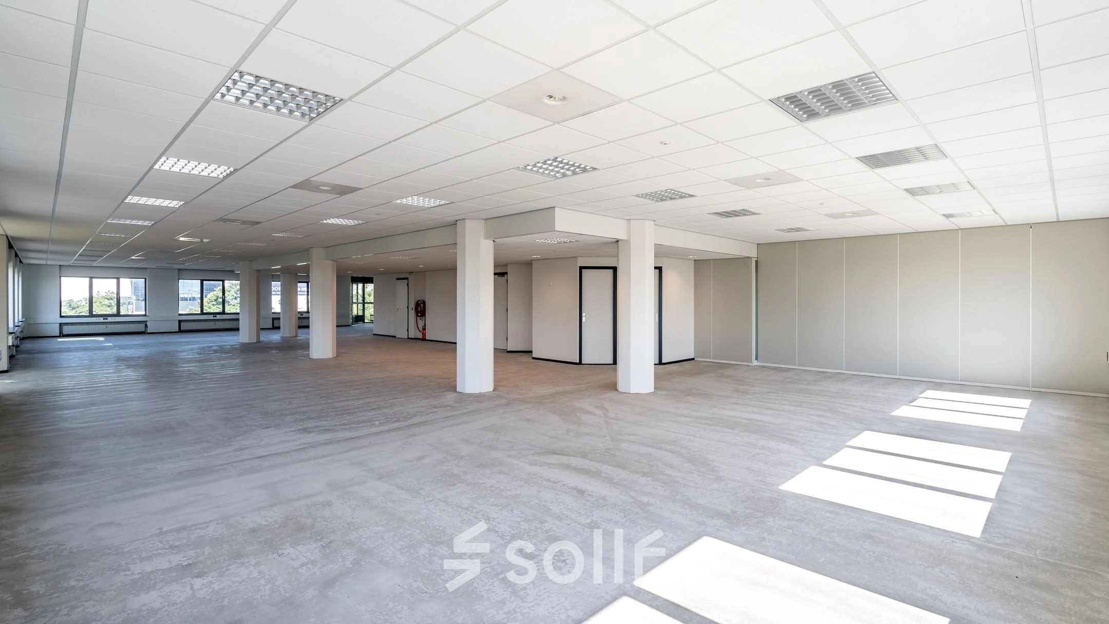 Open and spacious office interior in Schiedam with large windows allowing natural light, ideal for office space rental. Neutral grey flooring and white walls offer a versatile setup for businesses.