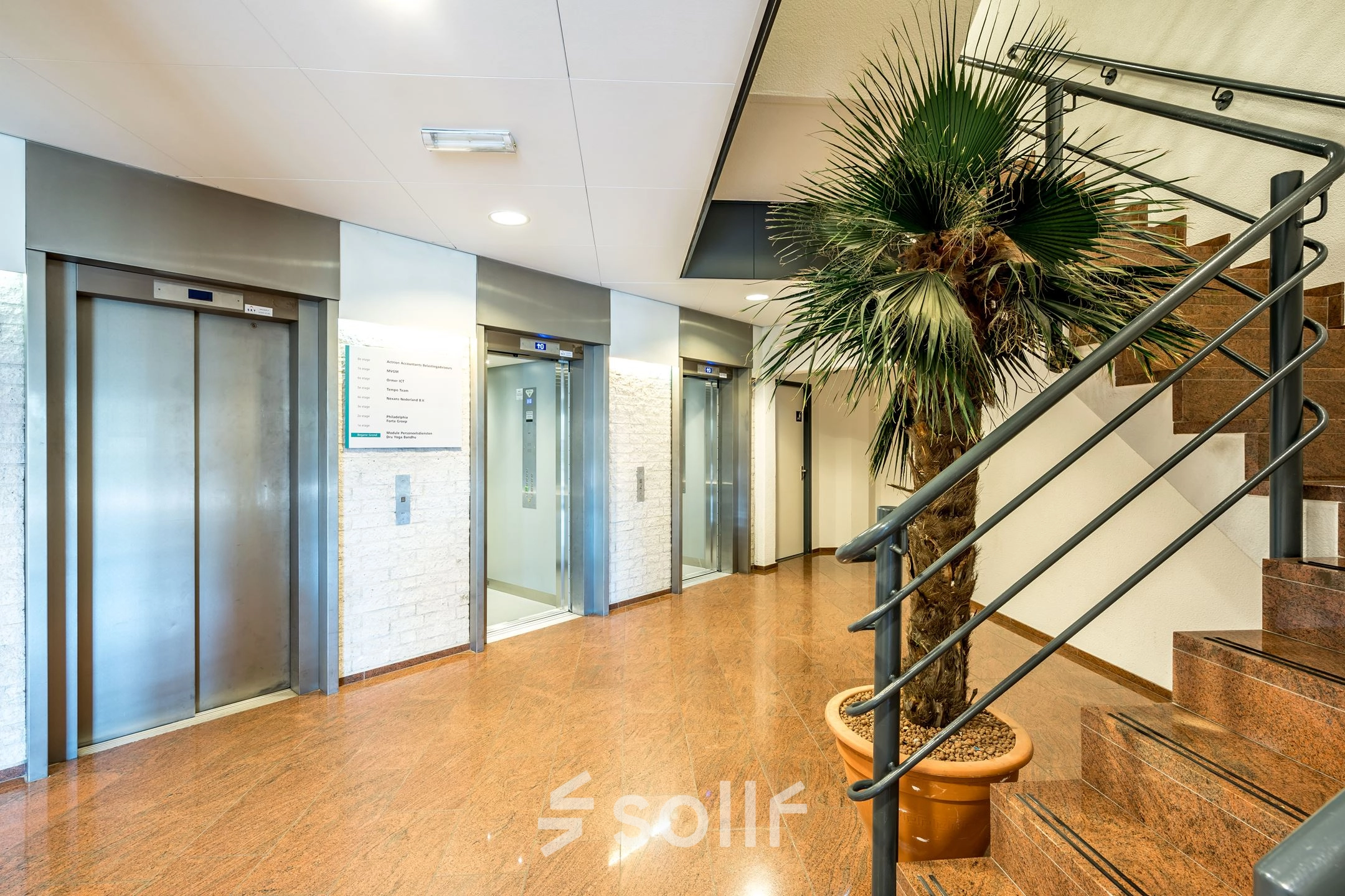 Interior view of the office to rent at Overschieseweg 304-332, Schiedam, featuring a sleek, polished floor, modern elevators, and a central staircase with a potted palm tree.