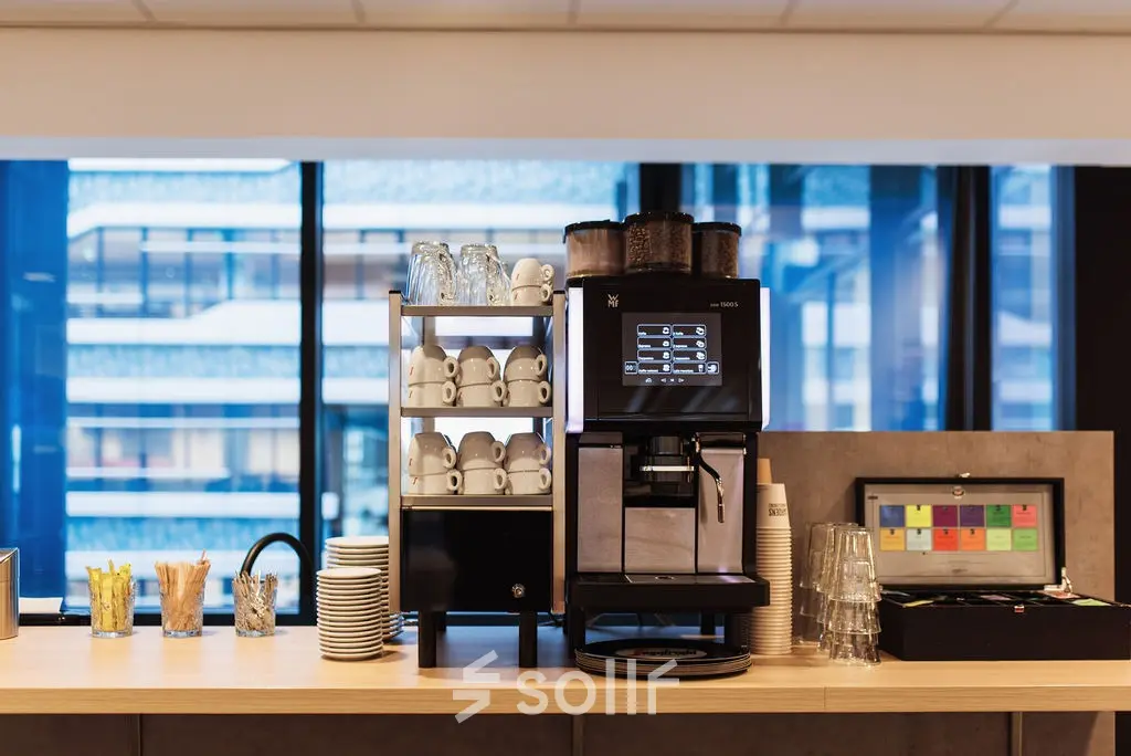 Office pantry area with a modern coffee machine, cups, and accessories set against a backdrop of large windows in The Hague Center. Ideal for office space rental.