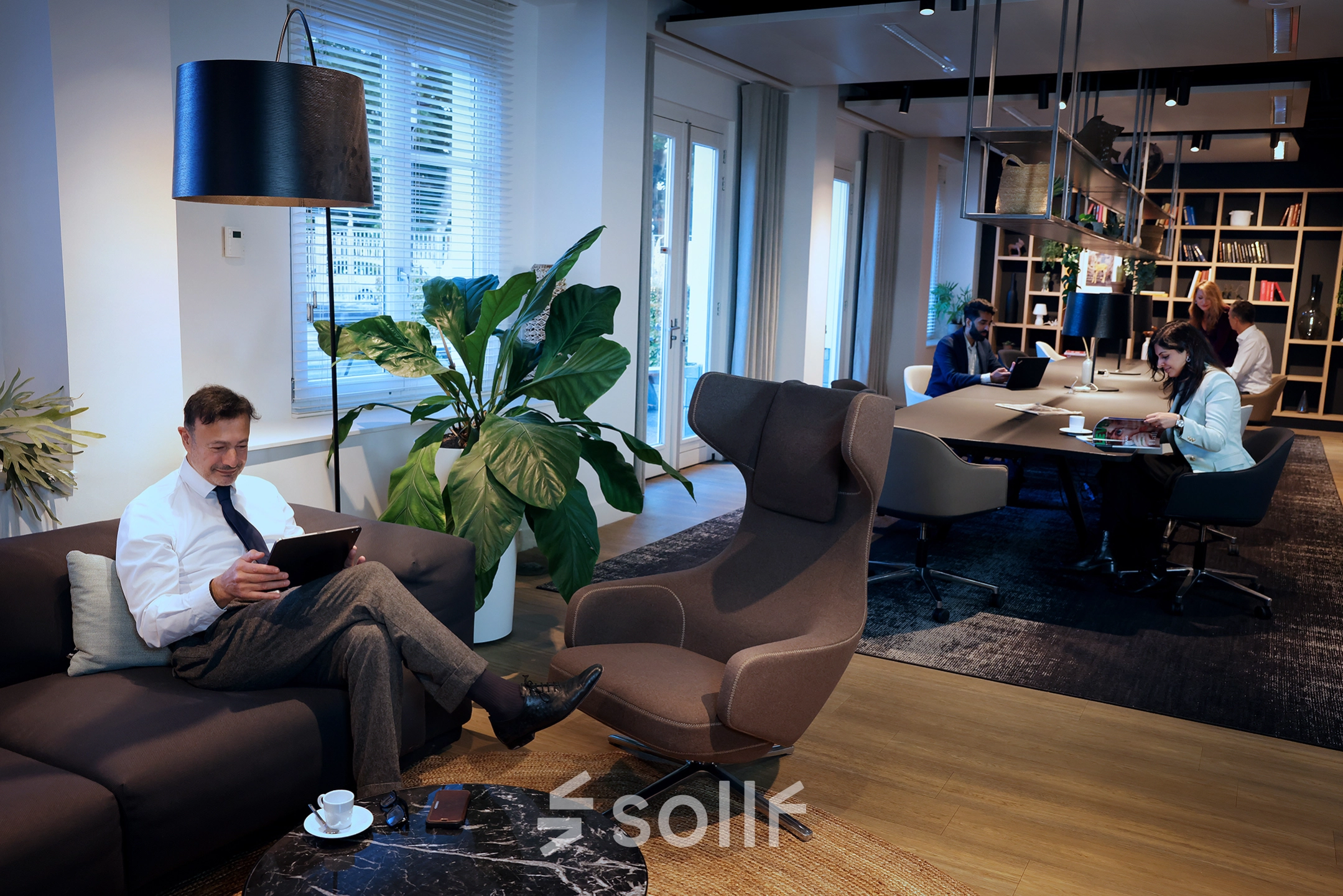 Modern office lounge with individuals working on laptops, featuring comfortable seating and stylish decor, located at Koninginnegracht 19 in The Hague Center, available for office space rental.