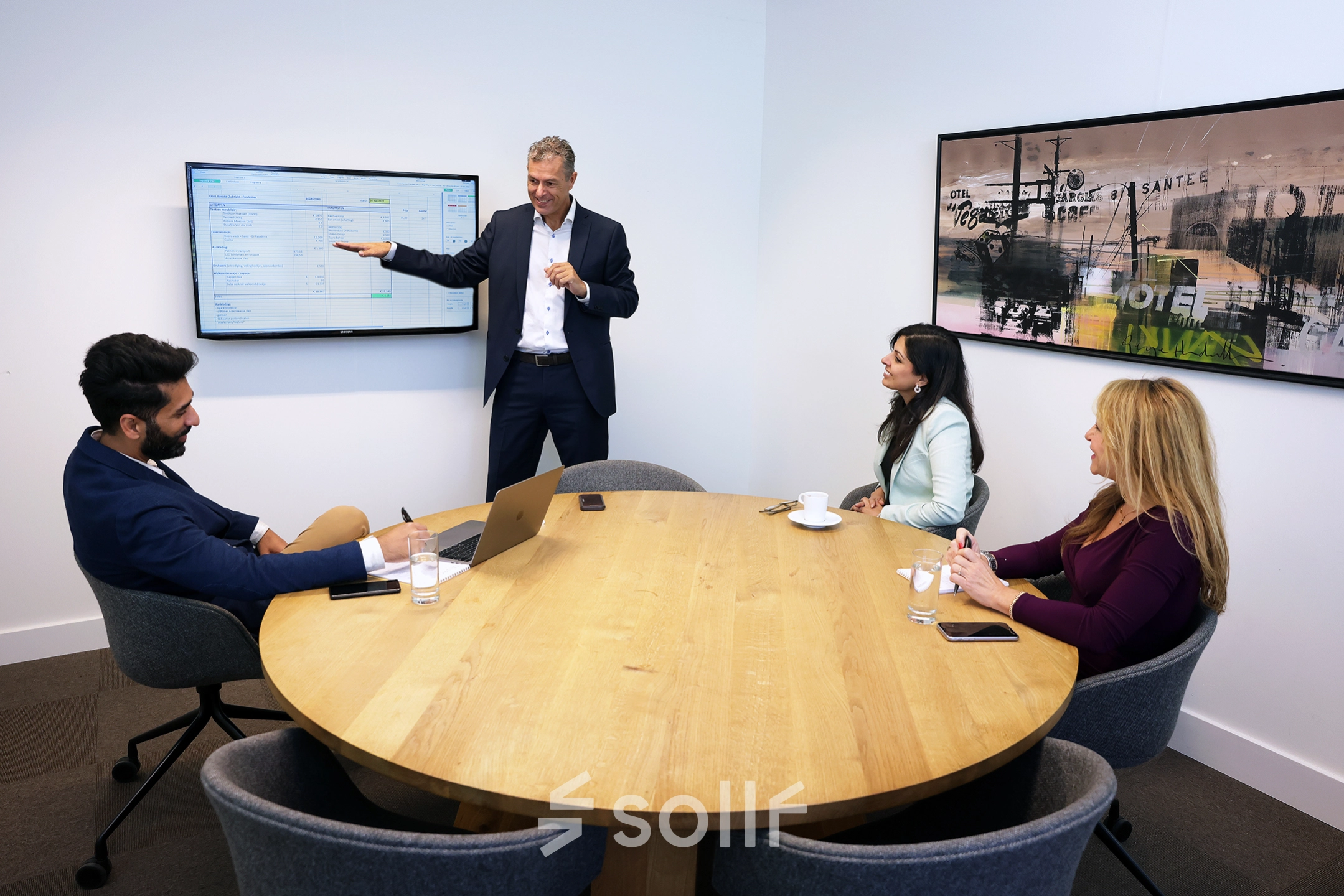 Professionals in a meeting around a wooden table in a furnished office, discussing information displayed on a wall-mounted screen at Koninginnegracht 19, ideal for office space rental.