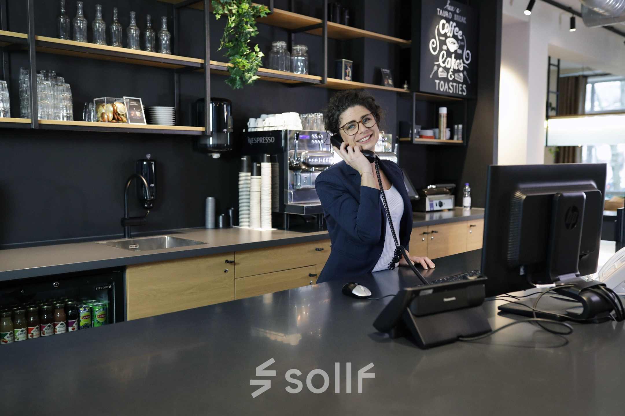 A reception area at President Kennedylaan 19 in The Hague Statenkwartier, featuring a smiling receptionist on the phone, modern furniture, and a well-stocked counter. Ideal office space rental.