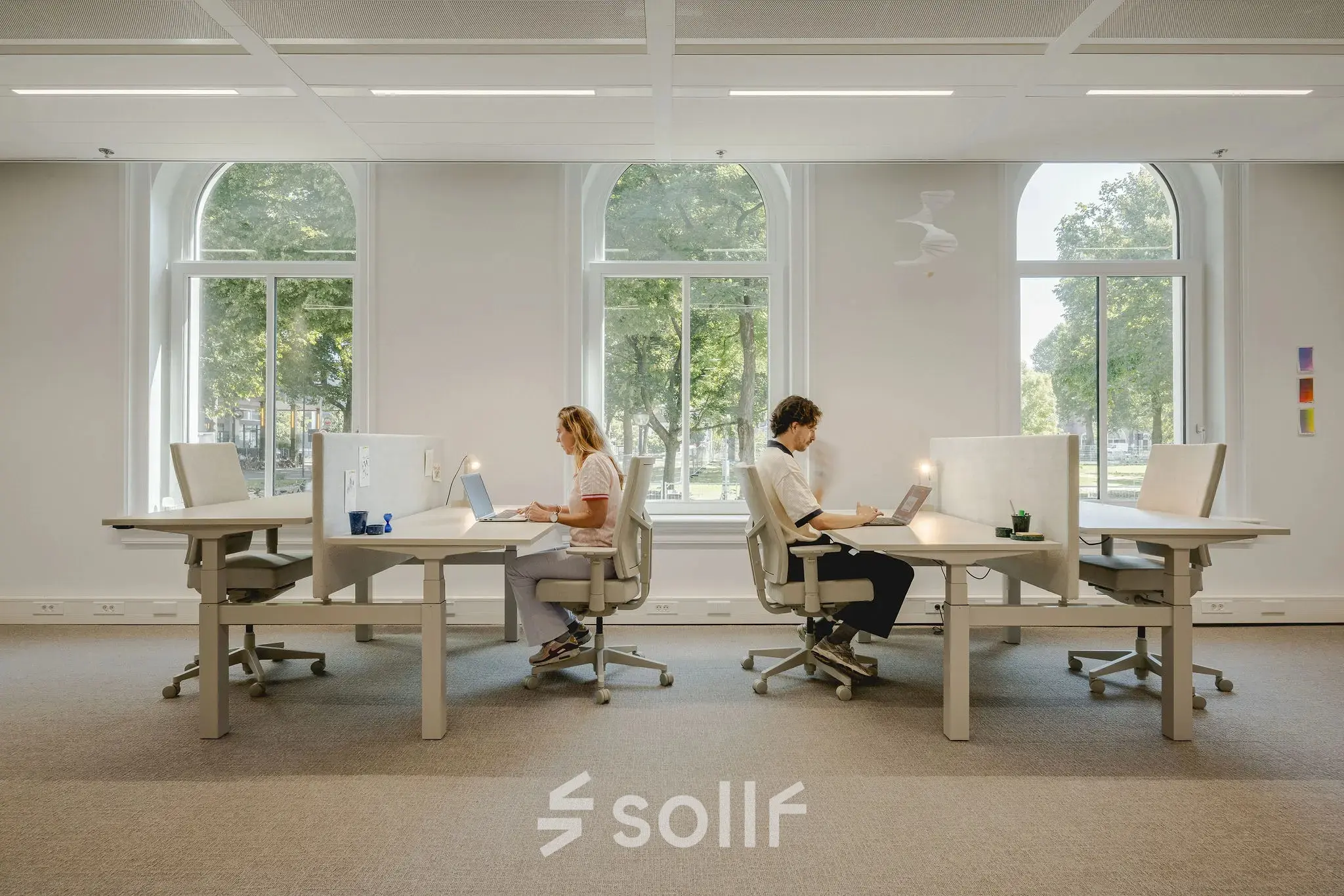 People working at desks in a bright, well-lit office space with large windows in Utrecht Central Station, ideal for office space rental.