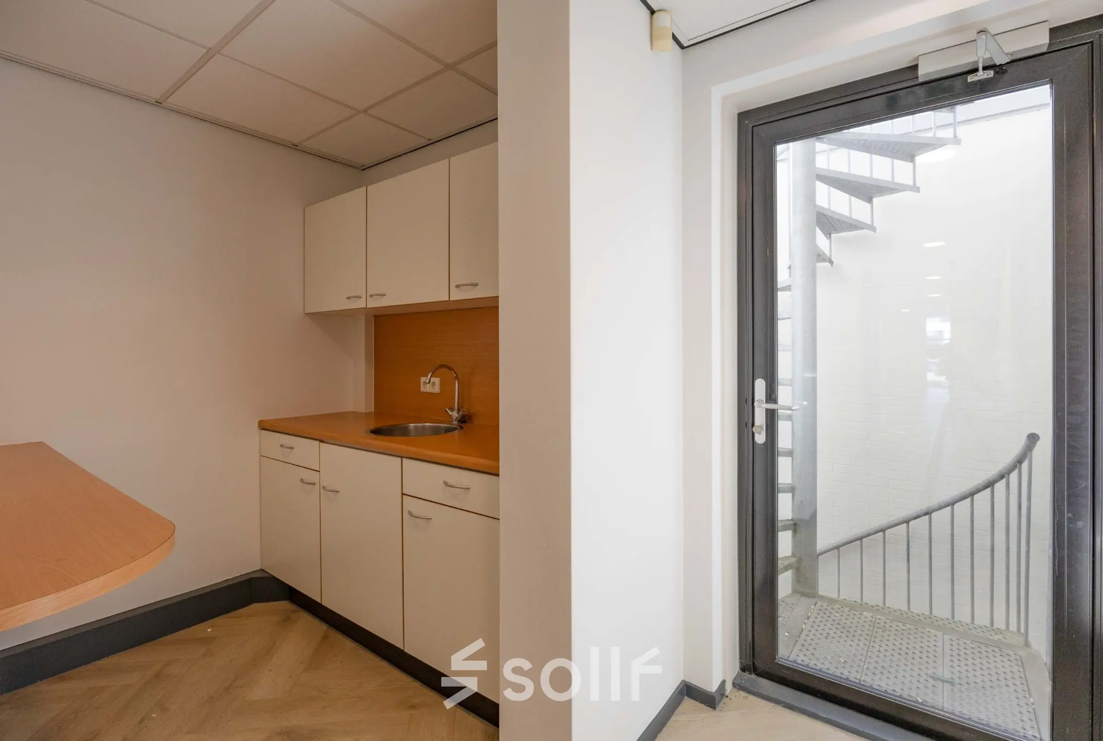 Simple office pantry at Keizersveld 47 in Venray, available for rent; features a small kitchenette with cabinets and sink next to a glass door leading outside.