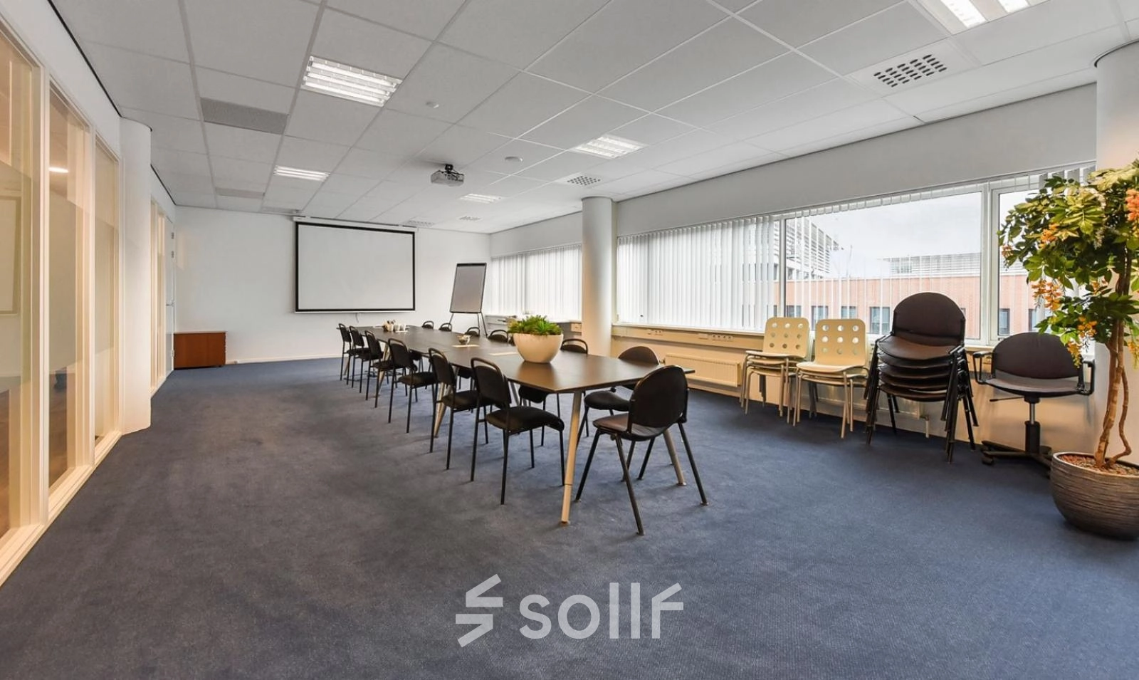Spacious furnished office space rental at Regulusweg 11, Voorburg, featuring a large conference room with a long table, chairs, potted plants, and a projector screen. Bright natural light enters through the windows.