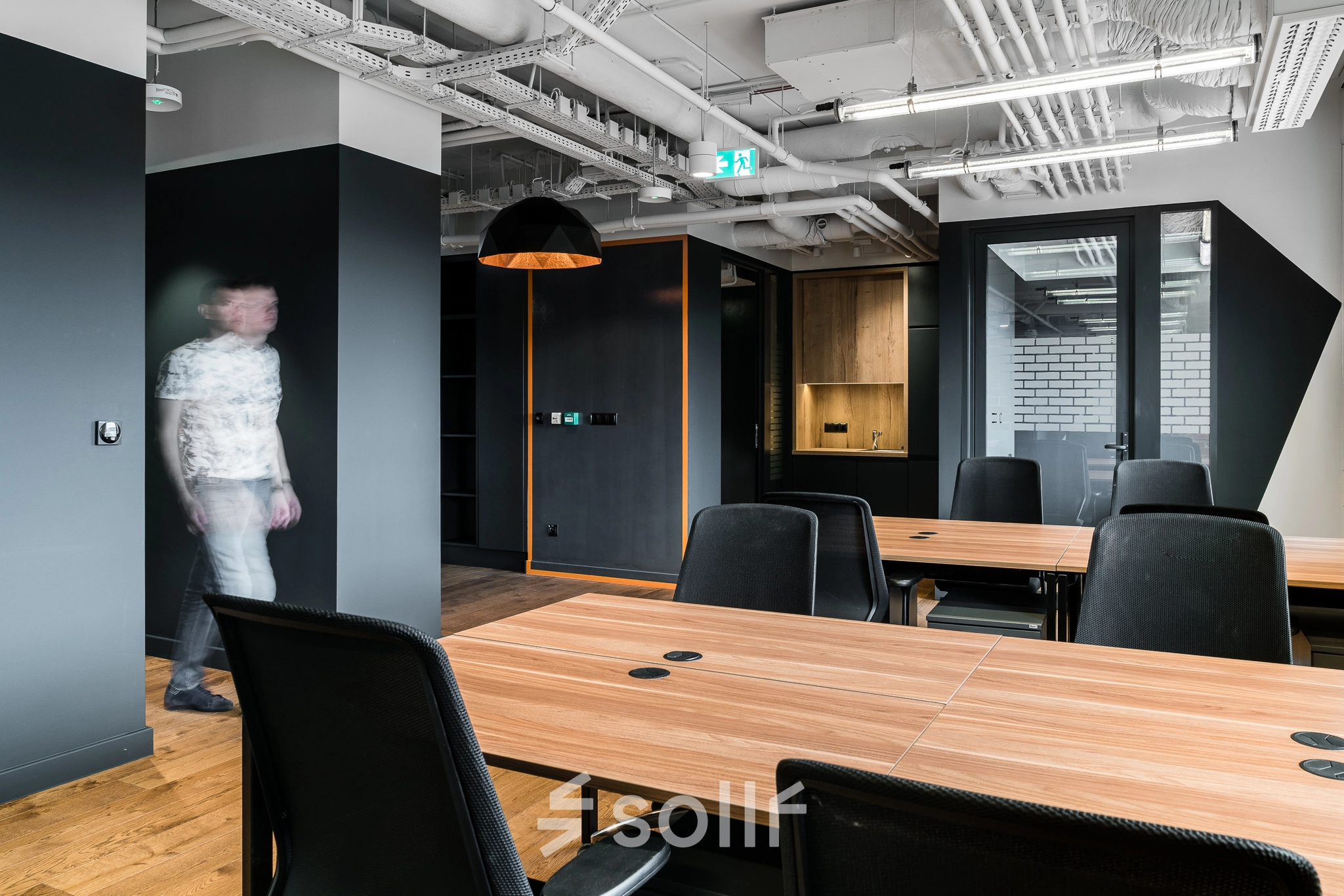 Modern office space with wooden desks and black chairs, featuring exposed ceilings and contemporary lighting at Konstruktorska 11, Warsaw Mokotow. A person walks through the workspace.
