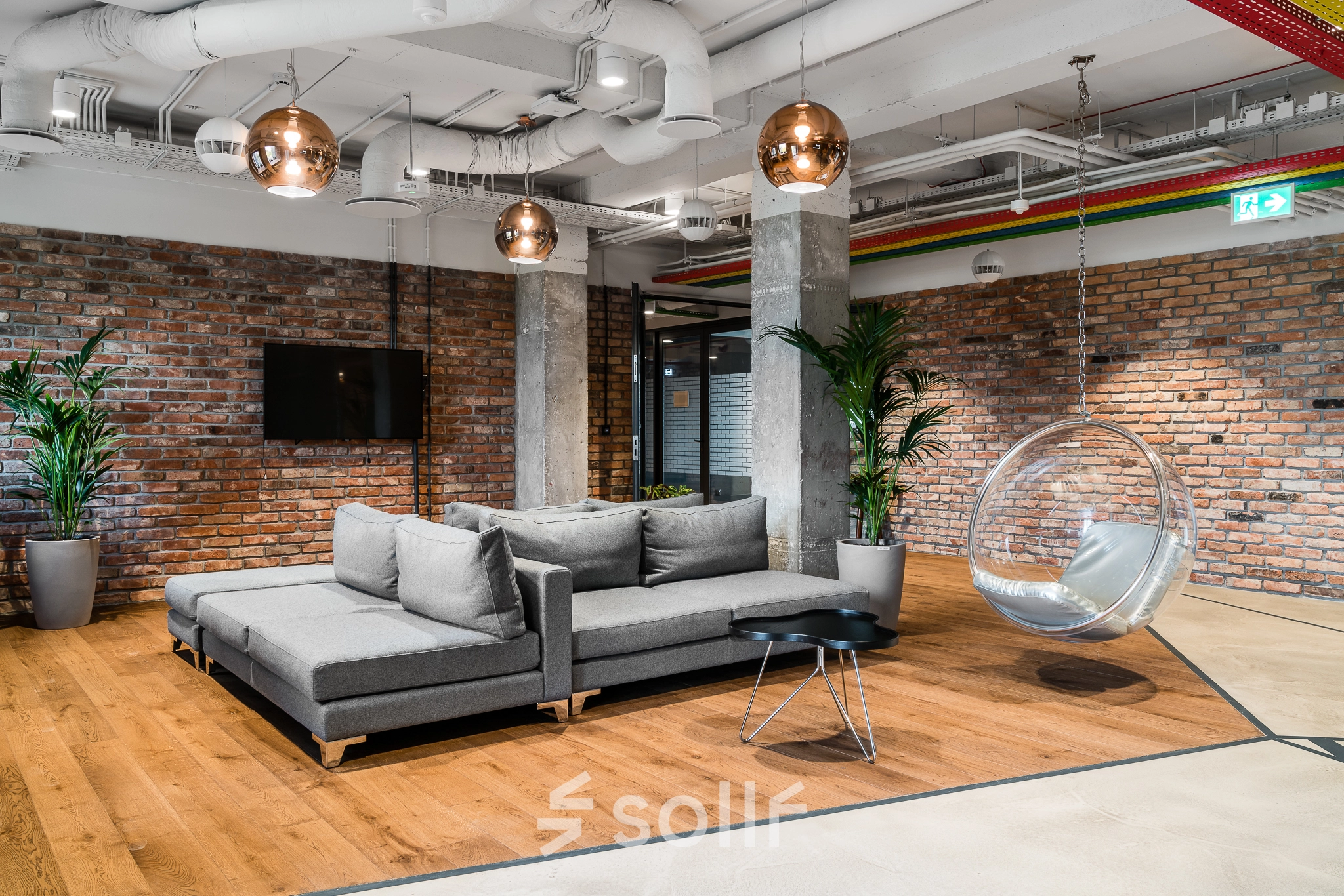 Modern office lounge at Konstruktorska 11, Warsaw Mokotow, with sleek furnishings, brick walls, and a hanging chair, available for office space rental.