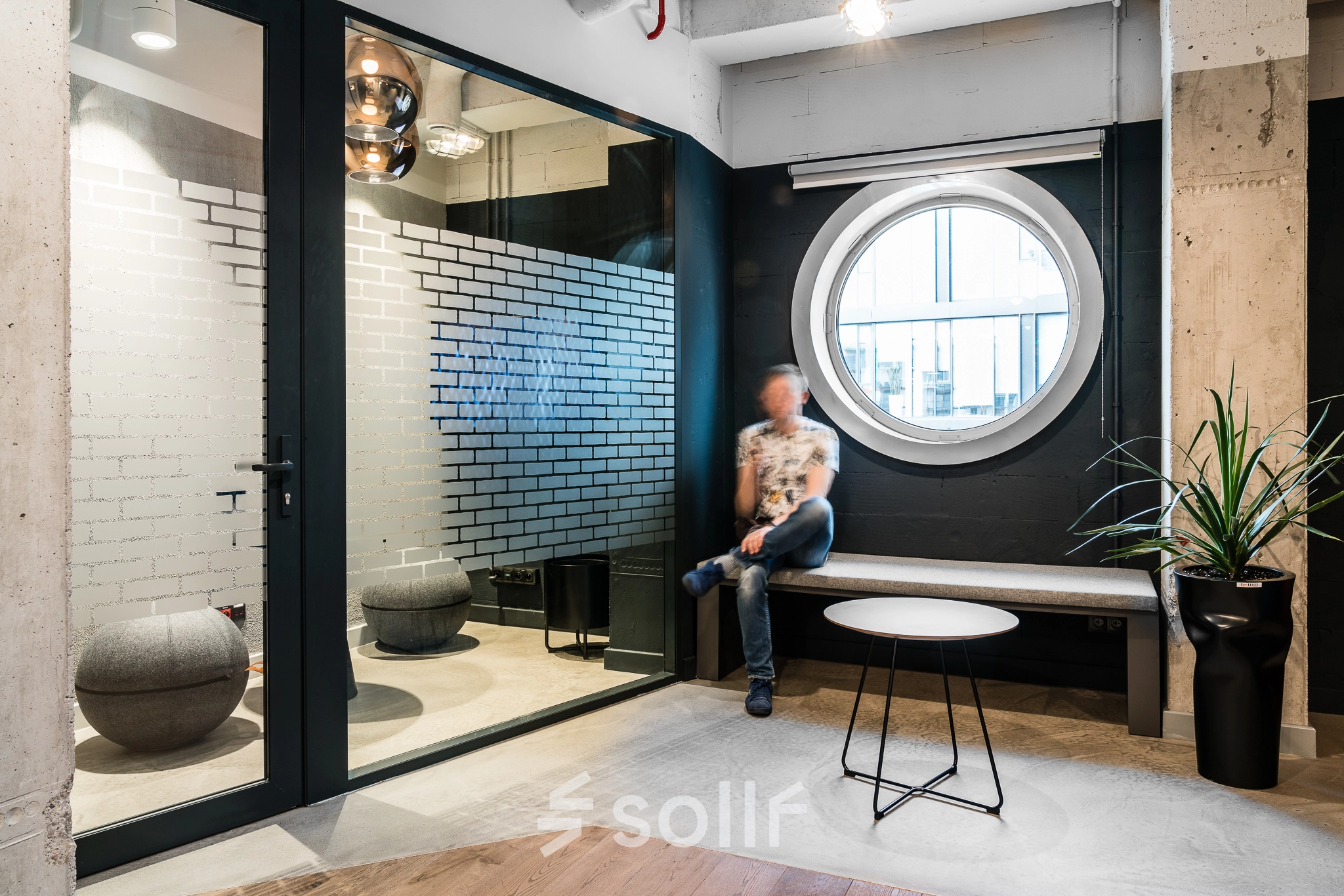 A cozy furnished office space with a round window and seating area at Konstruktorska 11, Warsaw Mokotow. A person is seated, engaging in light business activities.