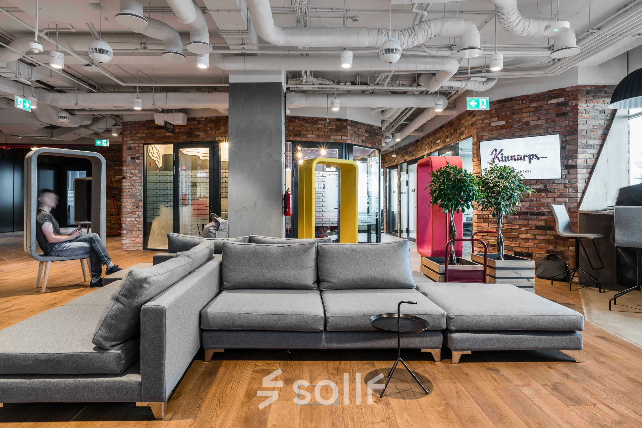 A modern furnished office on Konstruktorska 11, Warsaw Mokotow, featuring a gray sectional sofa and people engaged in conversation, highlighting an inviting space for office space rental.