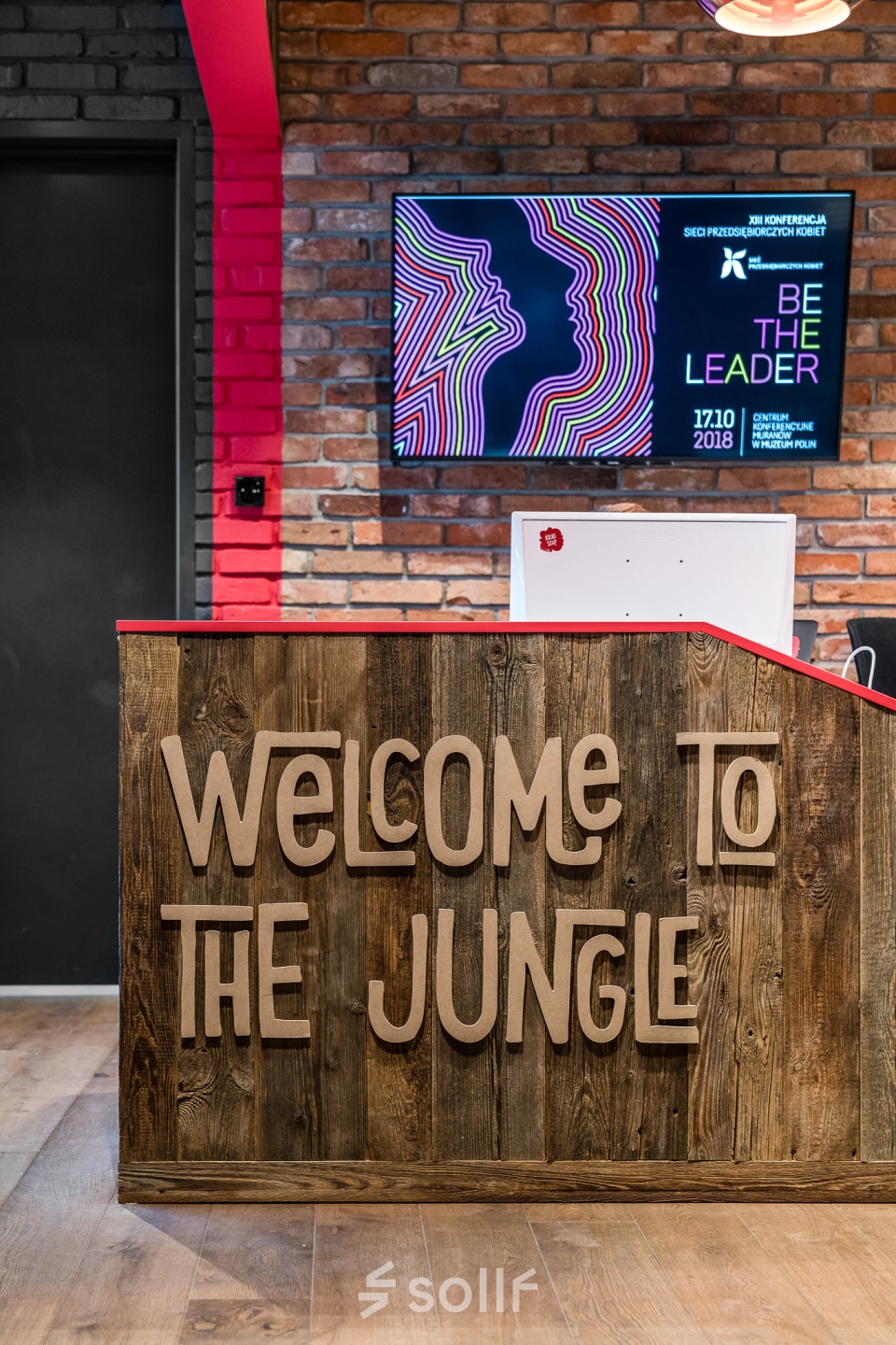 Reception area at Konstruktorska 11, Warsaw Mokotow, ideal for office space rental, featuring a wooden desk with 'Welcome to the Jungle' sign and a wall-mounted screen.