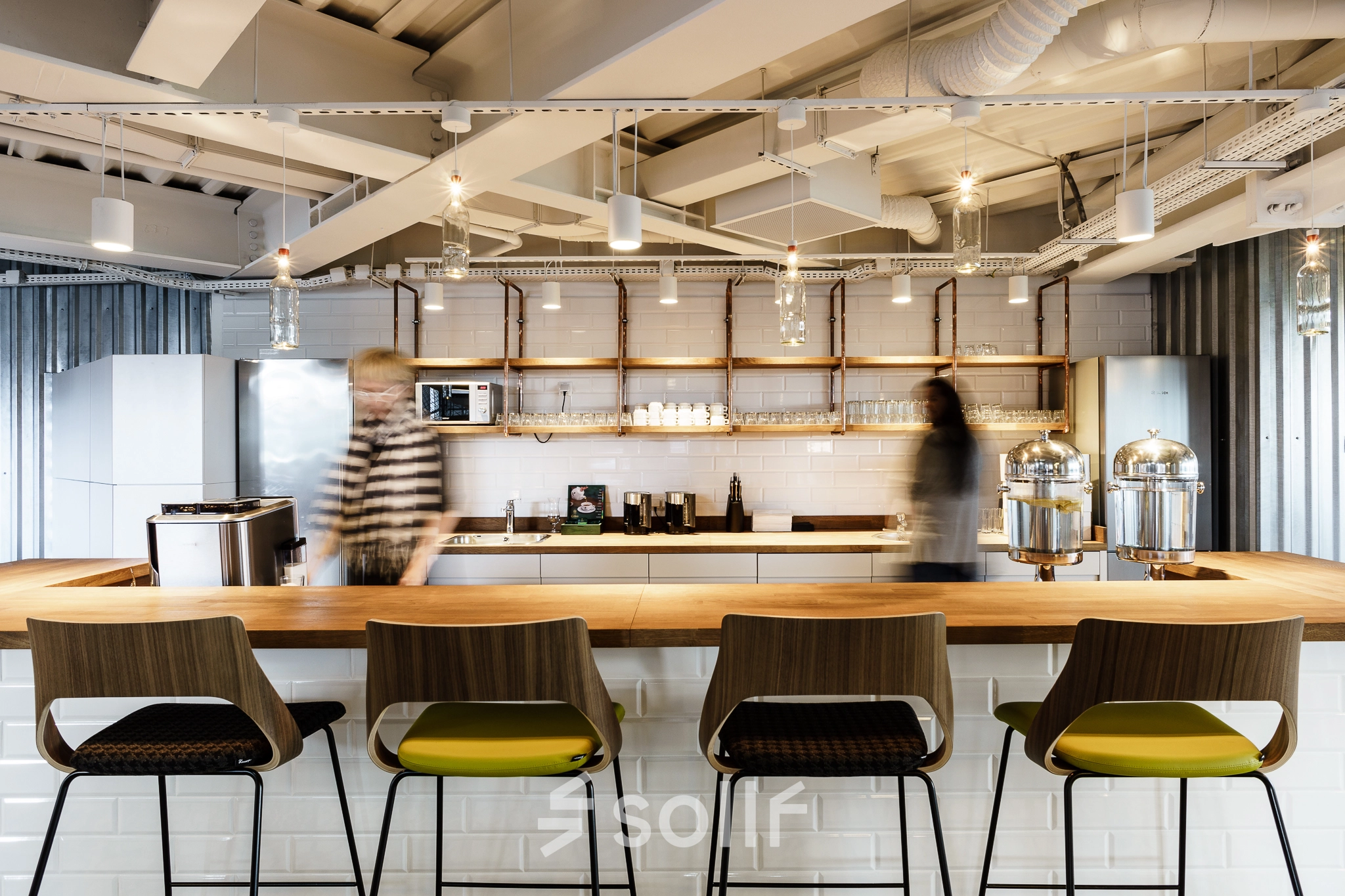 Modern office space in Warsaw Ochota, featuring a furnished kitchen area with sleek lighting and seating. Two individuals are engaging in work-related activities. Ideal for office space rental.
