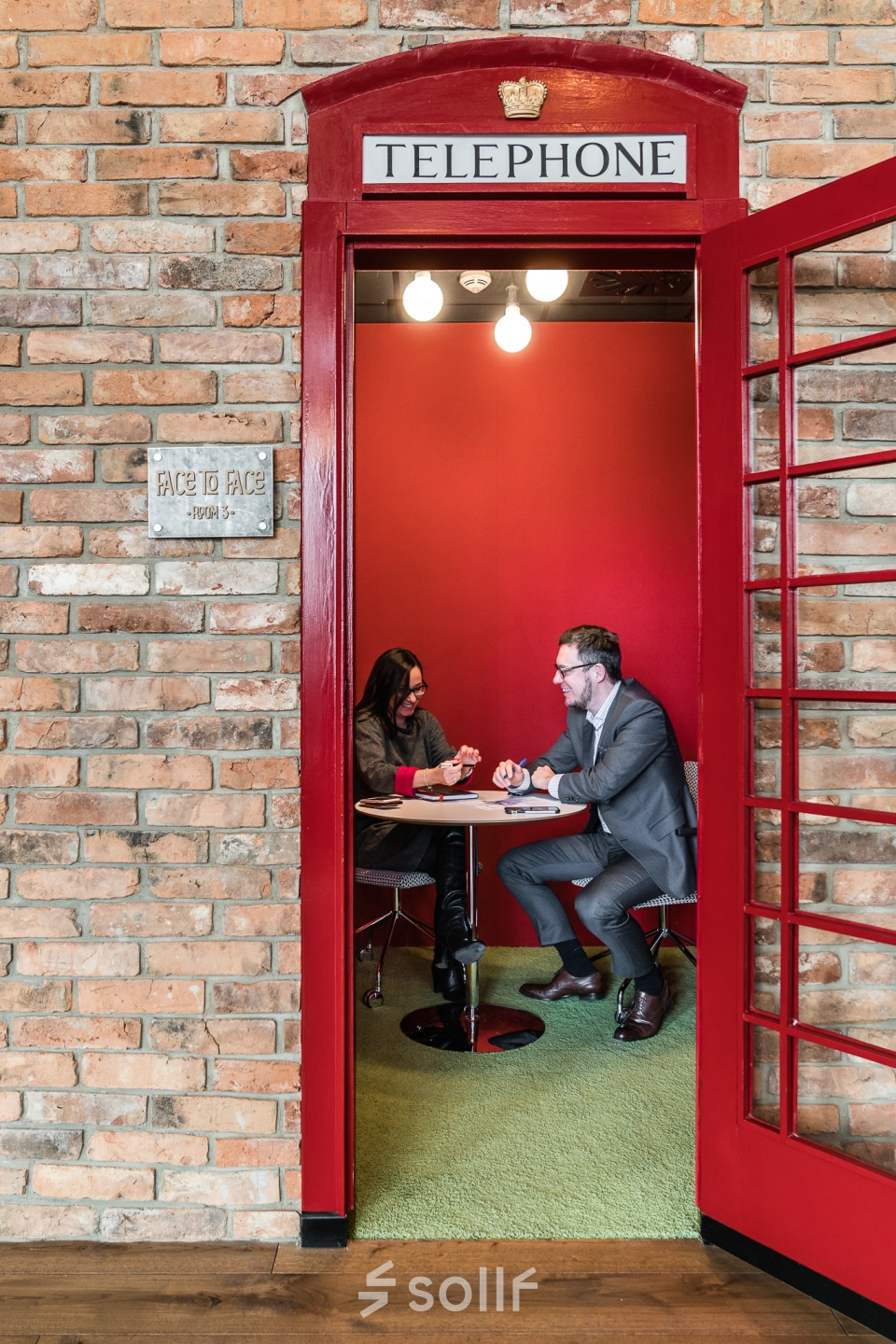 Two people are having a business meeting inside a unique telephone booth-style office space with red walls and a small table.