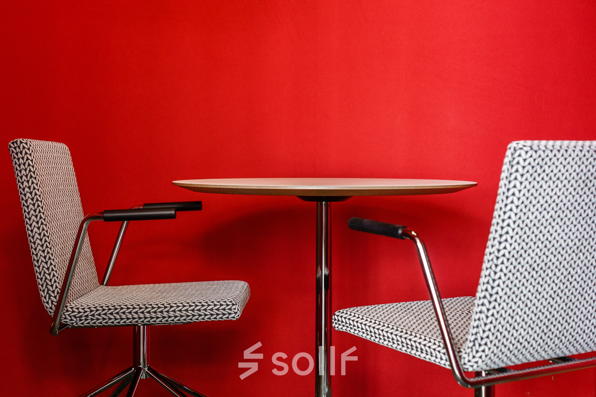 Furnished office space with two modern chairs and a small round table against a vibrant red wall, ideal for quick meetings or discussions.