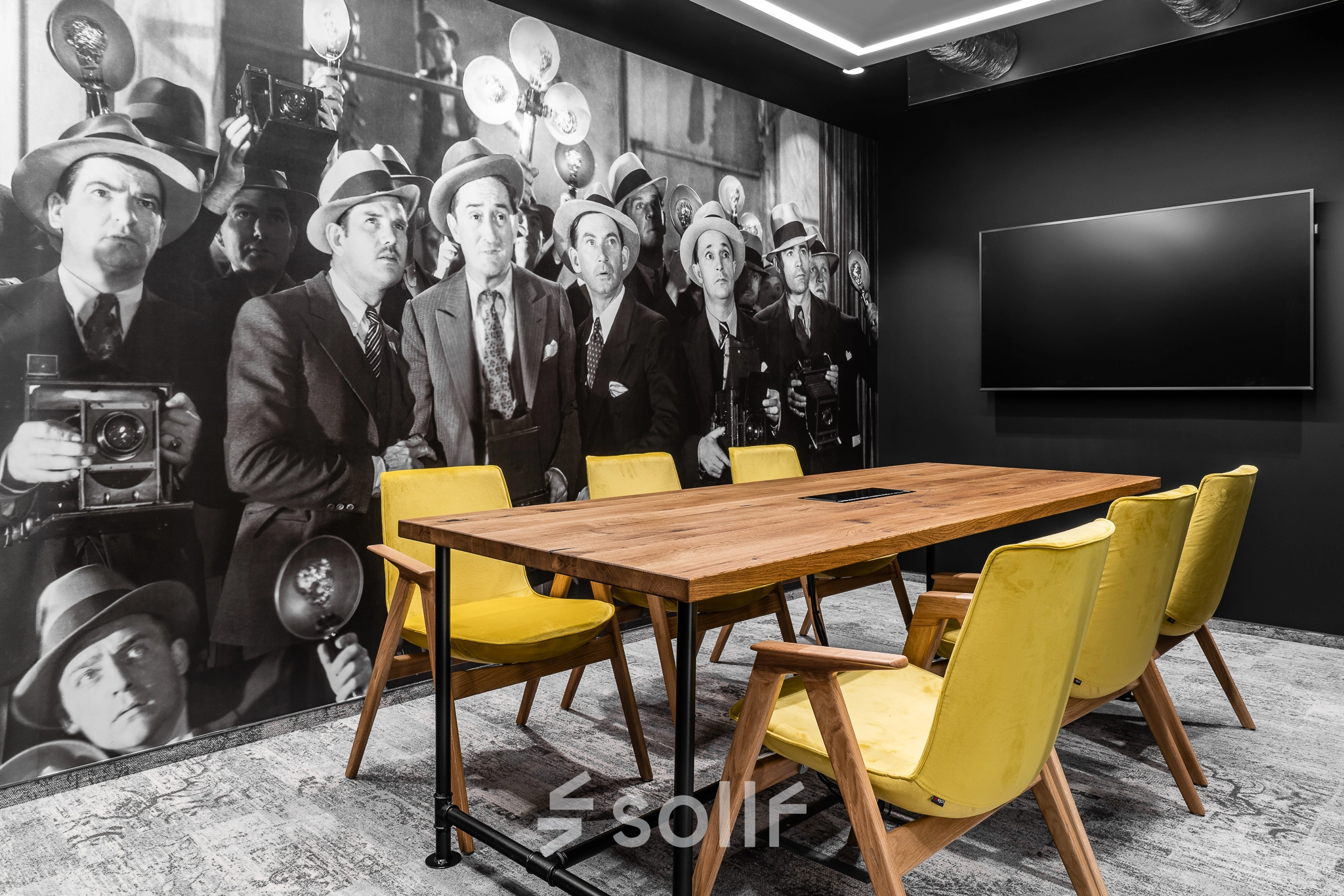 Furnished conference room with a large wooden table, yellow chairs, and a wall mural of photographers, ideal for office space rental.