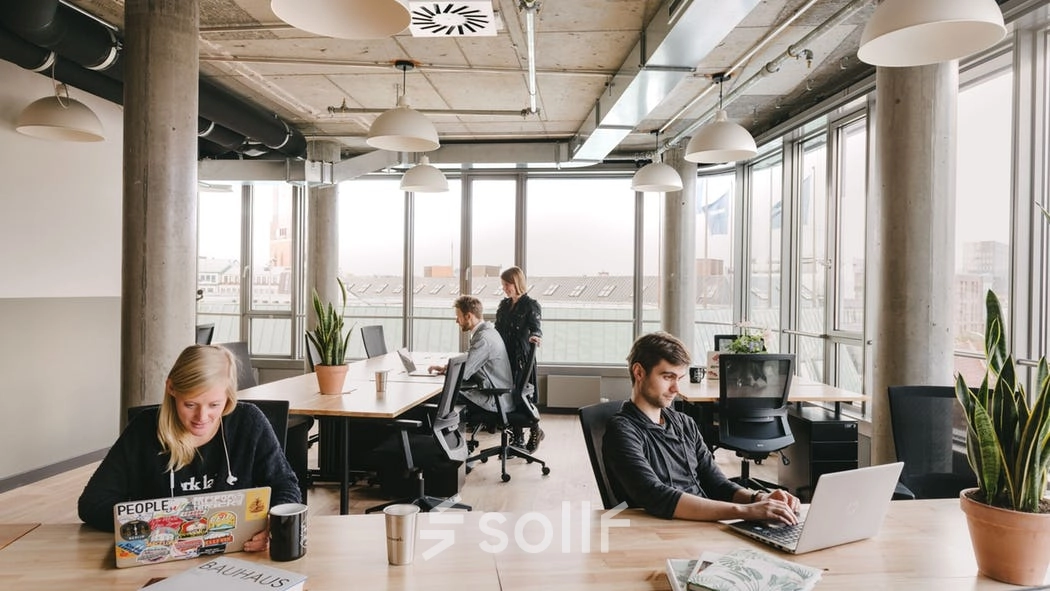 A modern office space with large windows at Grzybowska 60, Warsaw Wola, featuring employees working on laptops and holding discussions. Ideal for rent office space seekers.