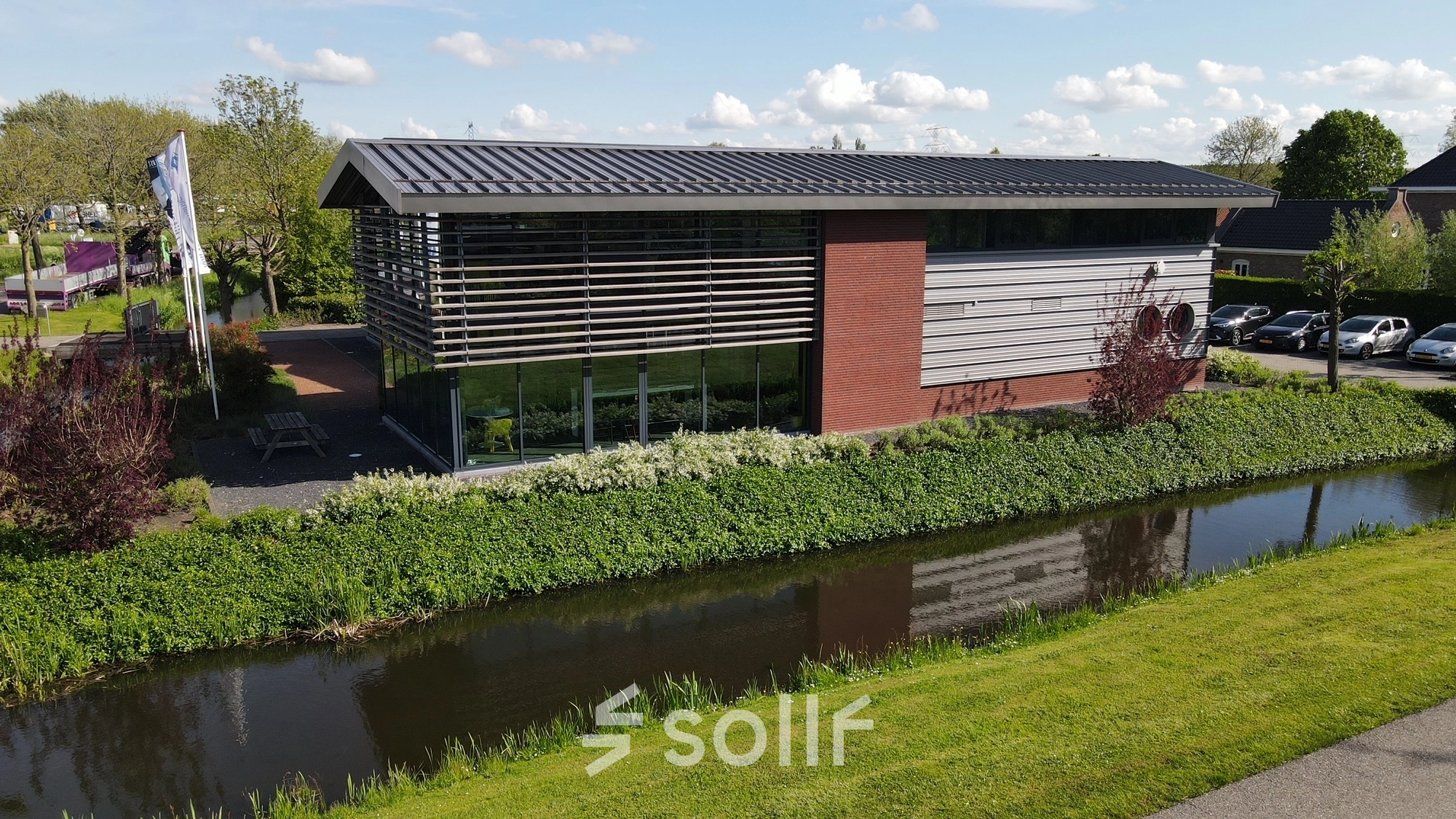 Exterior view of a modern office building for rent office space located at M.A. Reinaldaweg 2d, Woerden. The structure features sleek lines with large glass windows and a landscaped setting beside a canal.
