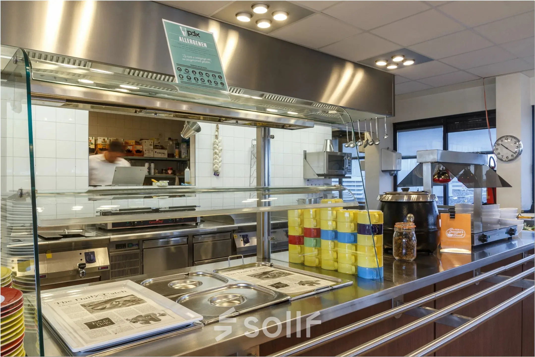A furnished office pantry with a serving counter, stacked plates, and utensils, located at Bredewater 26, Zoetermeer. An individual behind the counter prepares food.