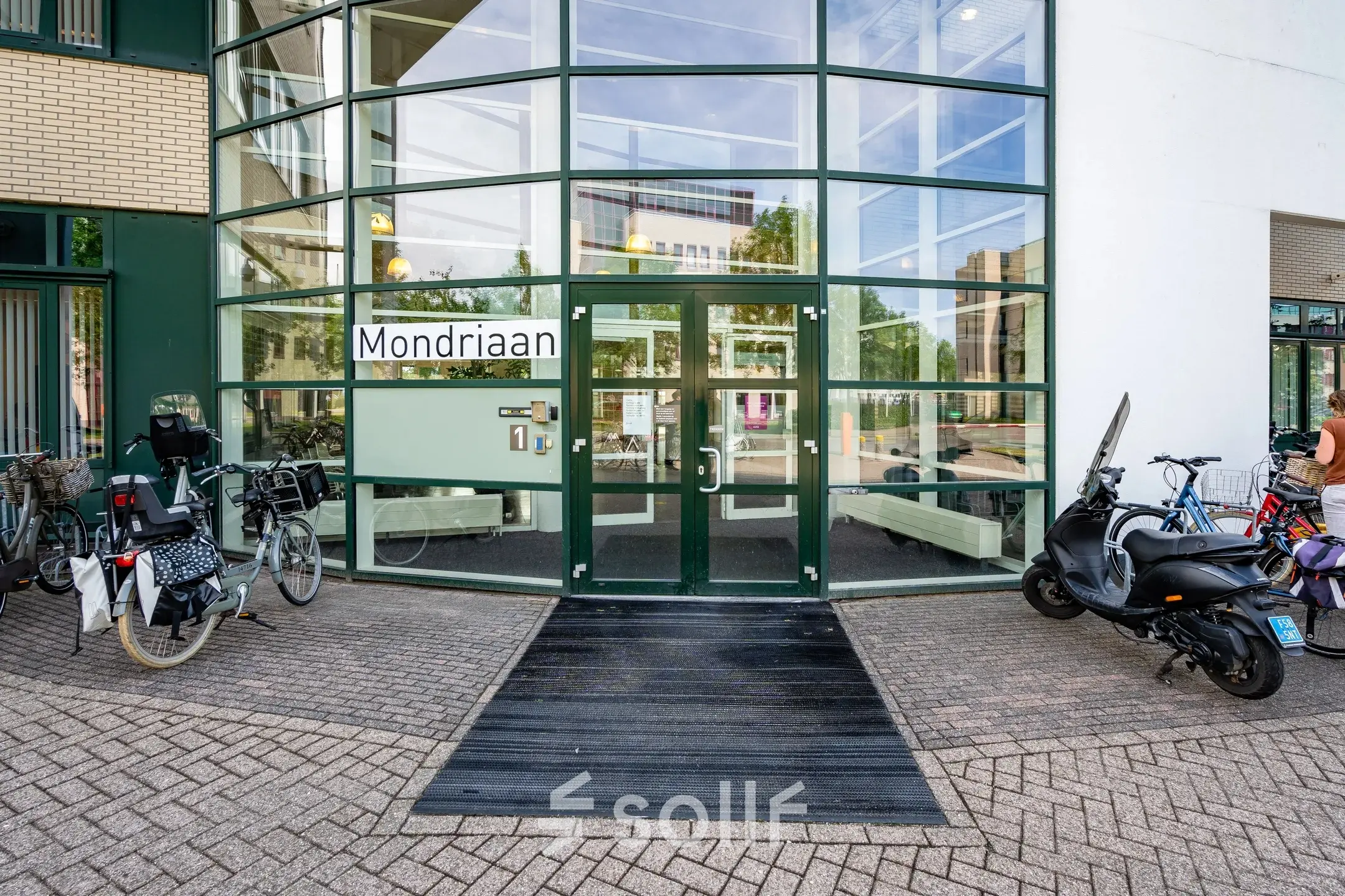 Exterior view of an office building at Dokter van Deenweg 1, Zwolle, highlighting the entrance with glass facades and a mix of bikes and scooters parked outside.
