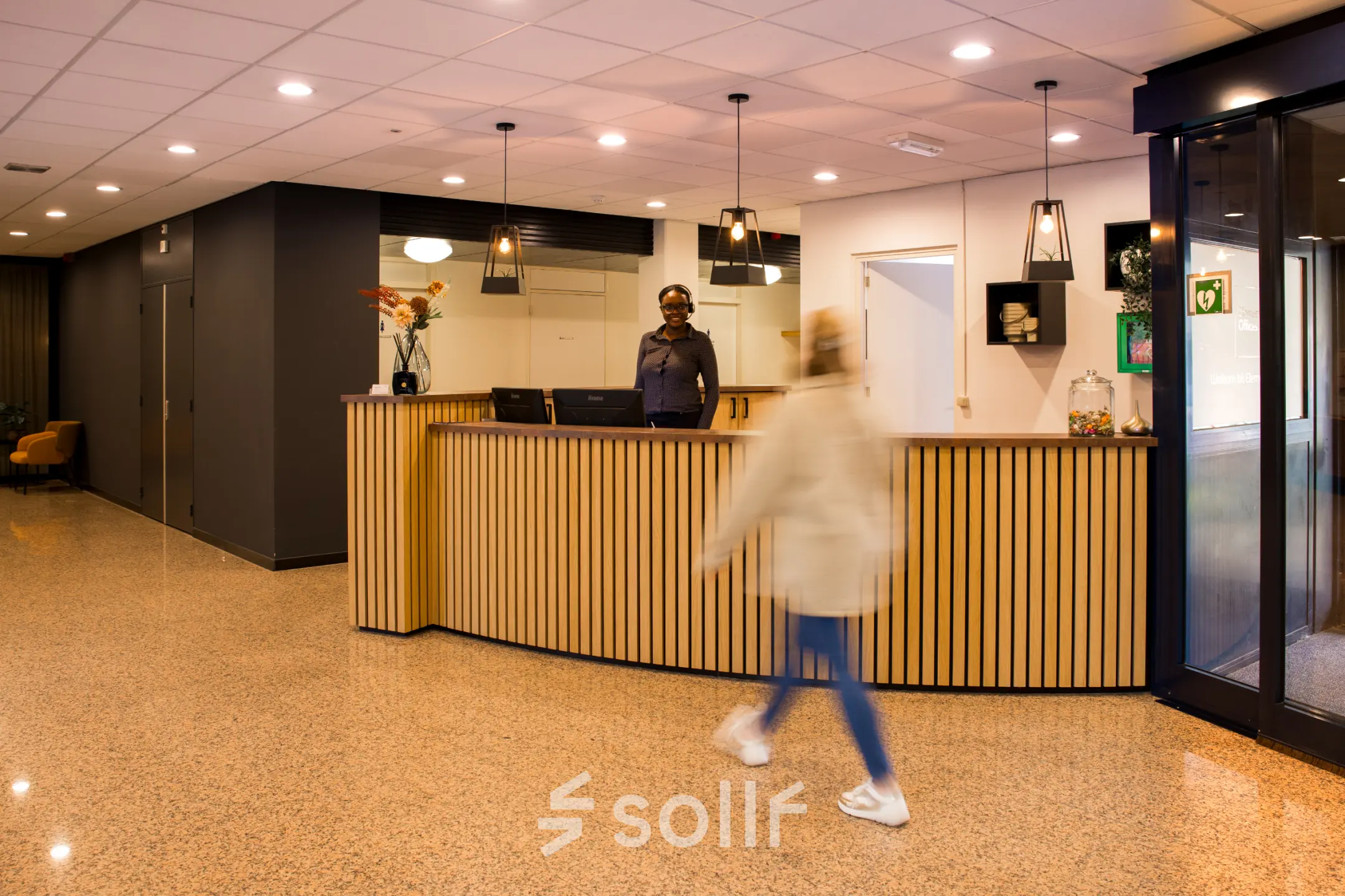 Kantoorruimte huren bij Markerkant 1310, Almere Stad. Moderne balie met verlicht plafond en houten details. Medewerker aan receptie, persoon loopt langs. Kantoorpand voor huur.