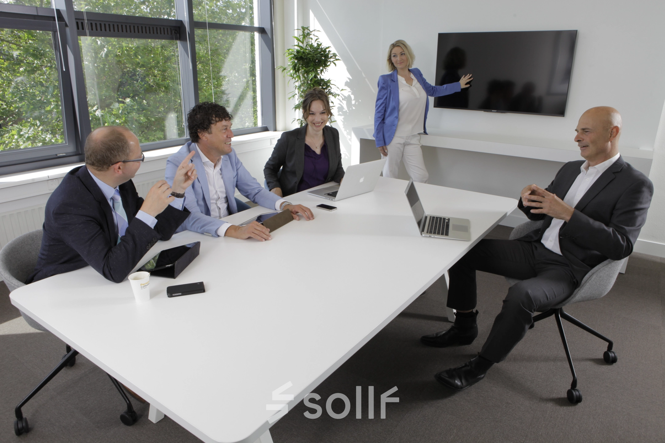 Zakelijke bijeenkomst in modern kantoorpand op Europalaan 16, Alphen aan den Rijn. Personen in bespreking rondom een witte tafel met laptops en scherm. Kantoorruimte huren mogelijk.