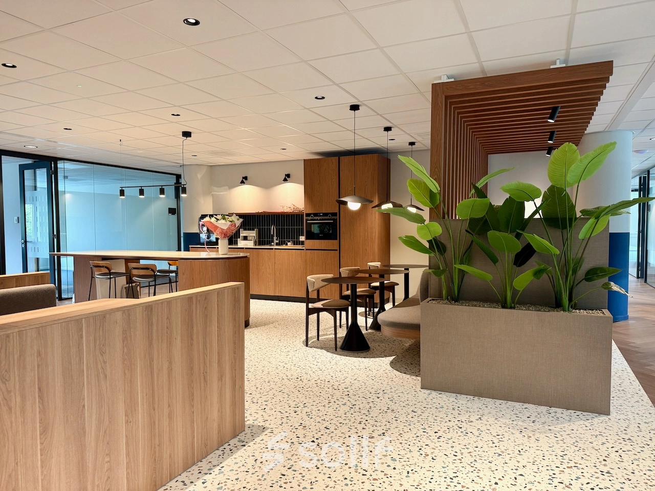 Moderne kantoorruimte in Amstelveen met houten en groene accenten, geschikt om te huren. Open werkruimte met stijlvolle meubels, planten en een kitchenette.