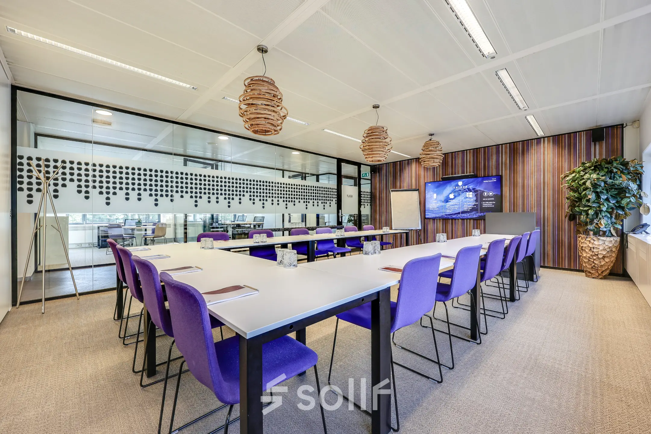 Vergaderruimte met paarse stoelen en grote tafels in een modern kantoorgebouw aan Prins Bernhardplein 200, Amsterdam Amstel. Ruime opzet met scherm en stijlvolle verlichting.