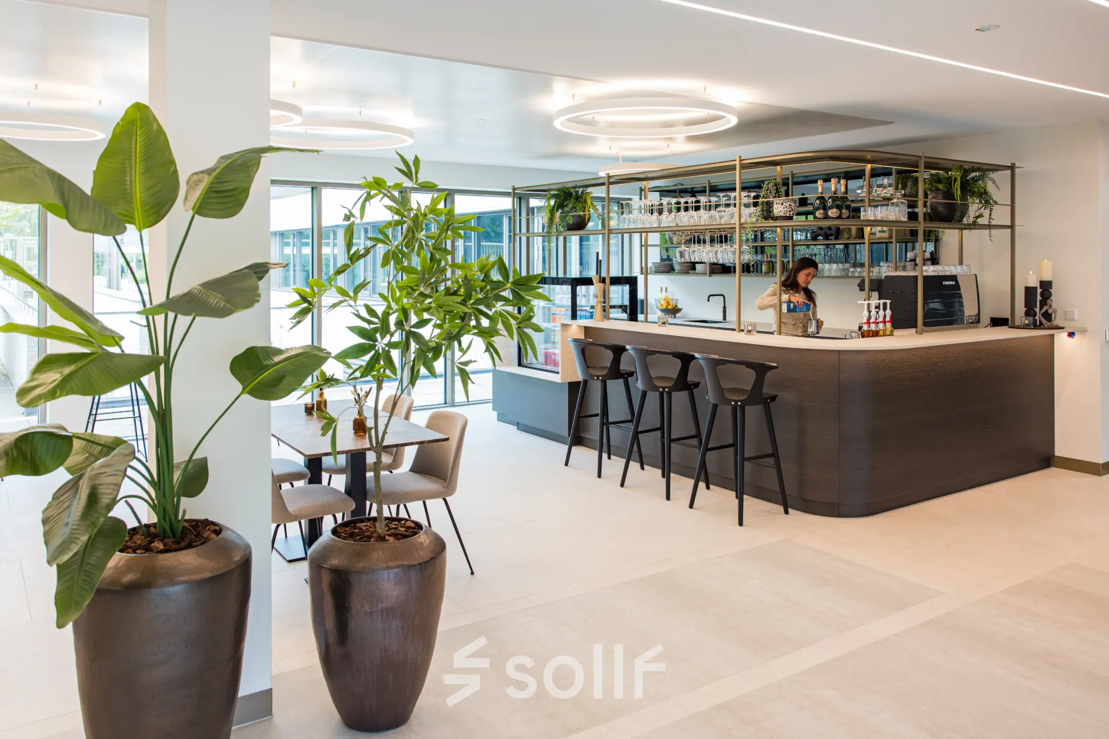 Moderne kantoorruimte huren op Walborg 2A in Amsterdam Buitenveldert, met een stijlvolle bar, grote planten en een vrouw die achter de balie bezig is.