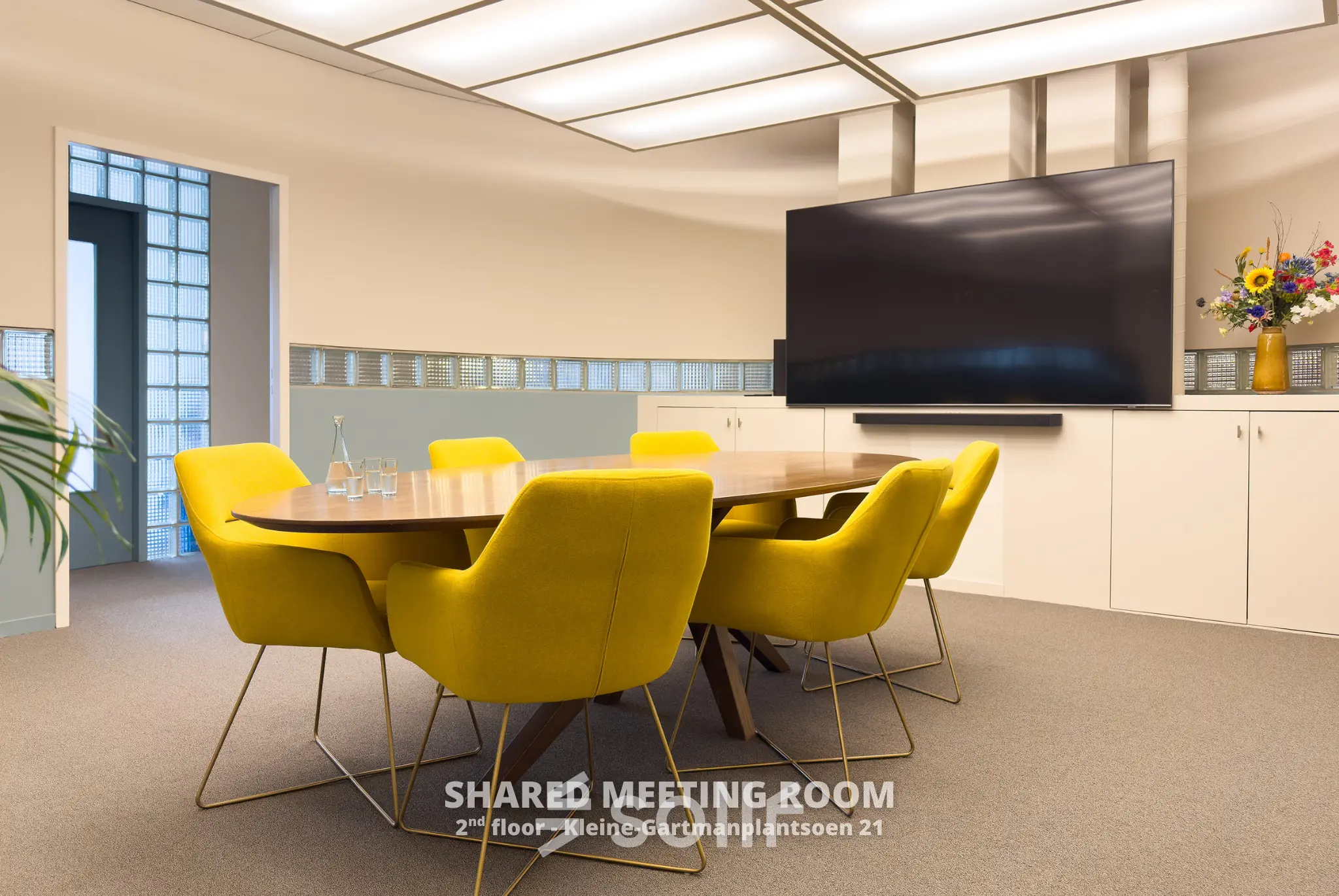 Vergaderruimte met felgele stoelen rond een houten tafel in het kantoorgebouw op Kleine Gartmanplantsoen 21, Amsterdam Centrum. Een televisie scherm aan de muur en een bloemstuk completeren de ruimte.