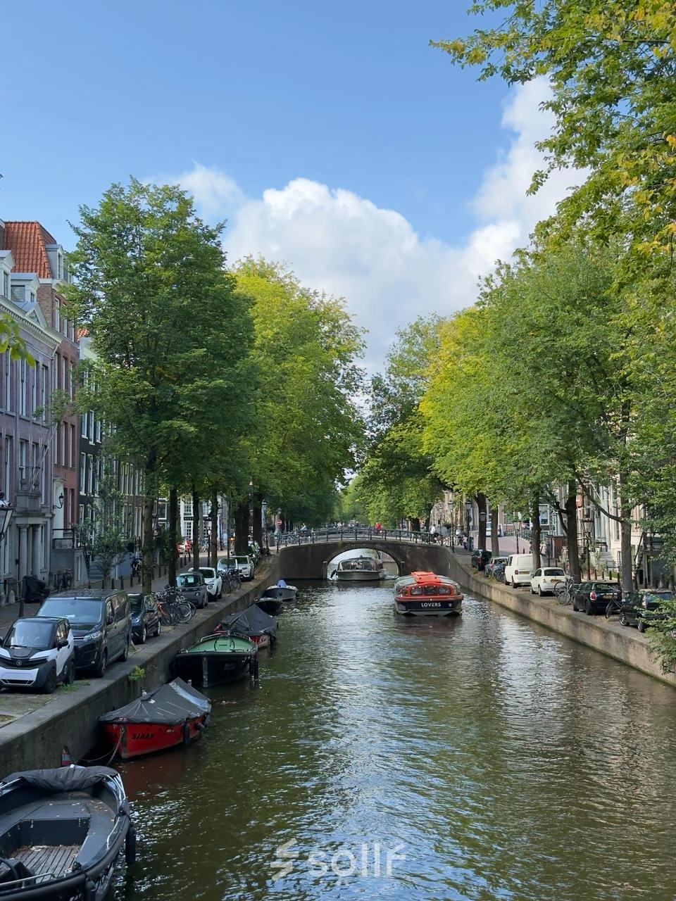 Uitzicht op de Amsterdamse gracht met boten, omringd door groene bomen langs Herengracht. Geen personen zichtbaar. Ideale locatie voor een kantoorruimte huren met authentieke charme.