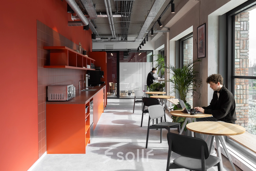 Moderne kantoorruimte huren in Amsterdam Noord met felrode keukenwand, houten tafels, en stoelen. Personen werken op laptops aan tafels bij grote ramen met veel natuurlijk licht.