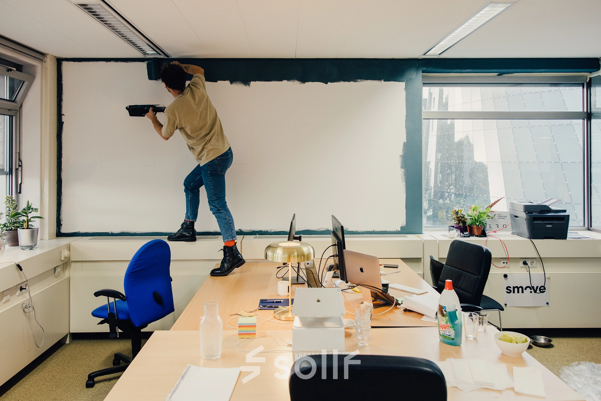 Een kantoor in Amsterdam Noord, TT Vasumweg 44, met een persoon die een muur schildert en bureaus die laptops, kantoorspullen en planten bevatten.