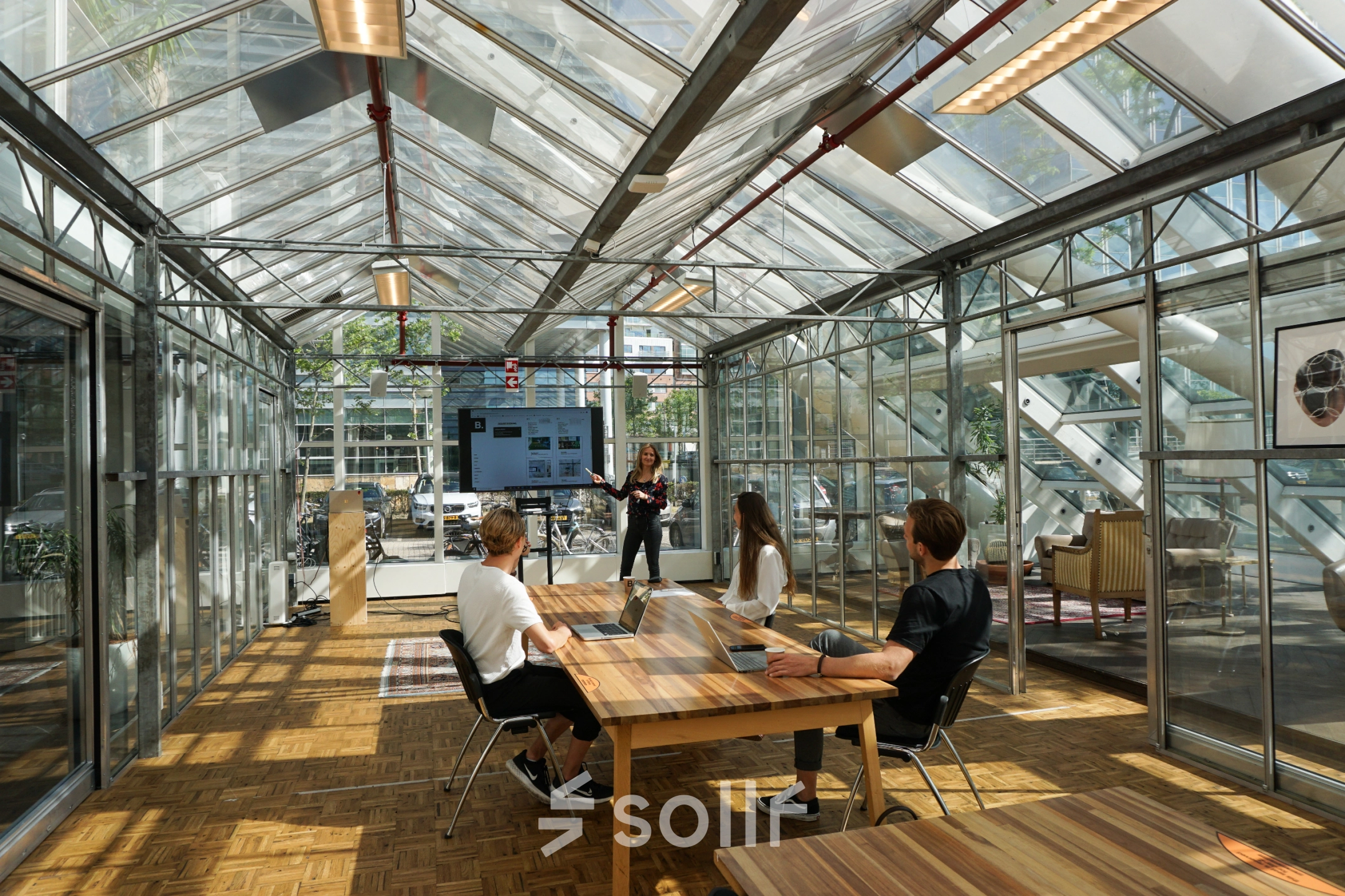 Kantoor met glazen wanden in Amsterdam Riekerpolder, gelegen op Johan Huizingalaan 400. Drie mensen bij vergadertafel, één presenteert aan een scherm. Ruimte biedt een heldere en moderne werkomgeving.
