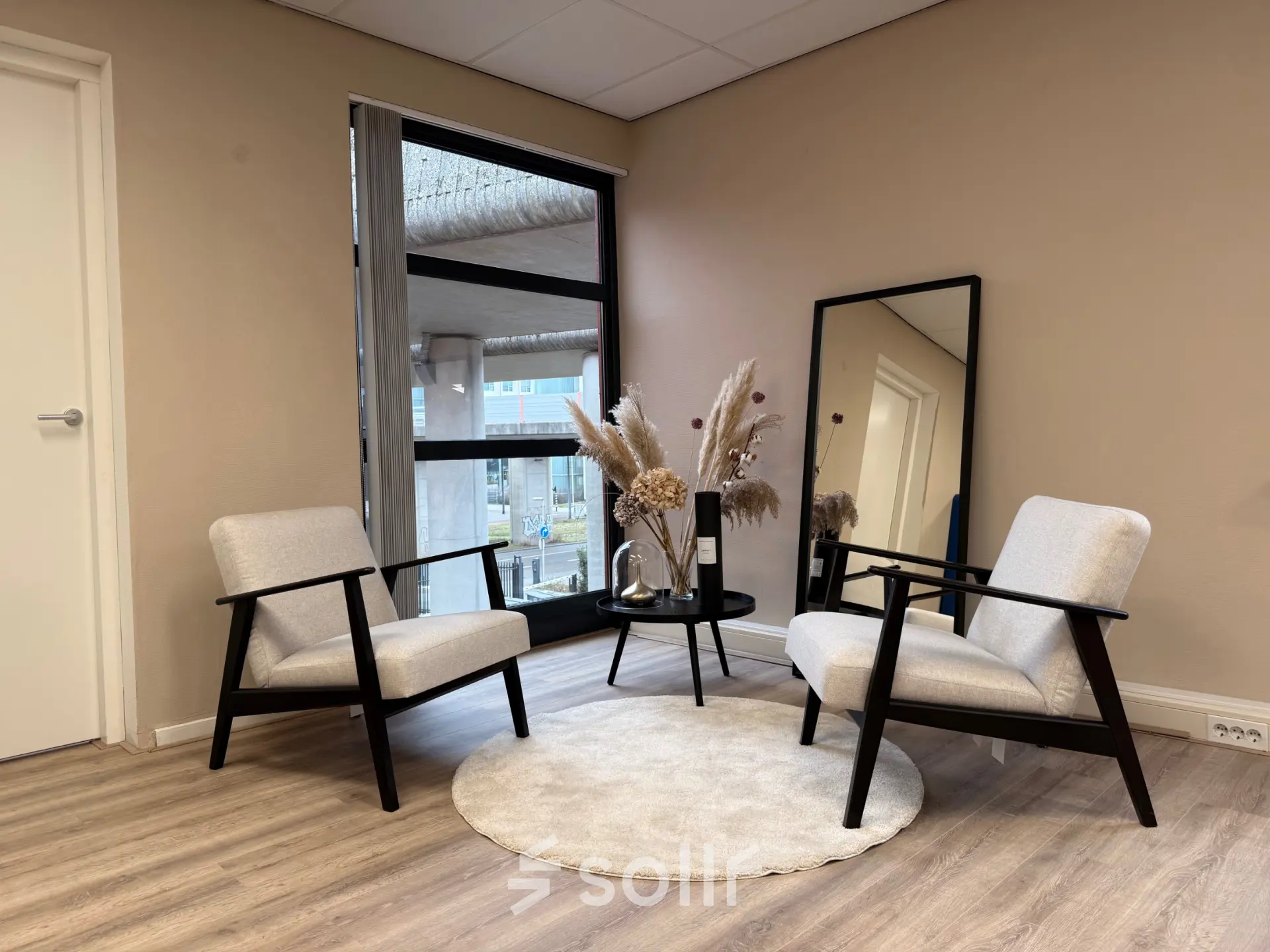 Kantoorinterieur op Arlandaweg 92, Amsterdam Sloterdijk, met een stijlvolle zithoek met twee stoelen, een rond kleed, en een bijzettafel. Grote ramen zorgen voor veel natuurlijk licht.