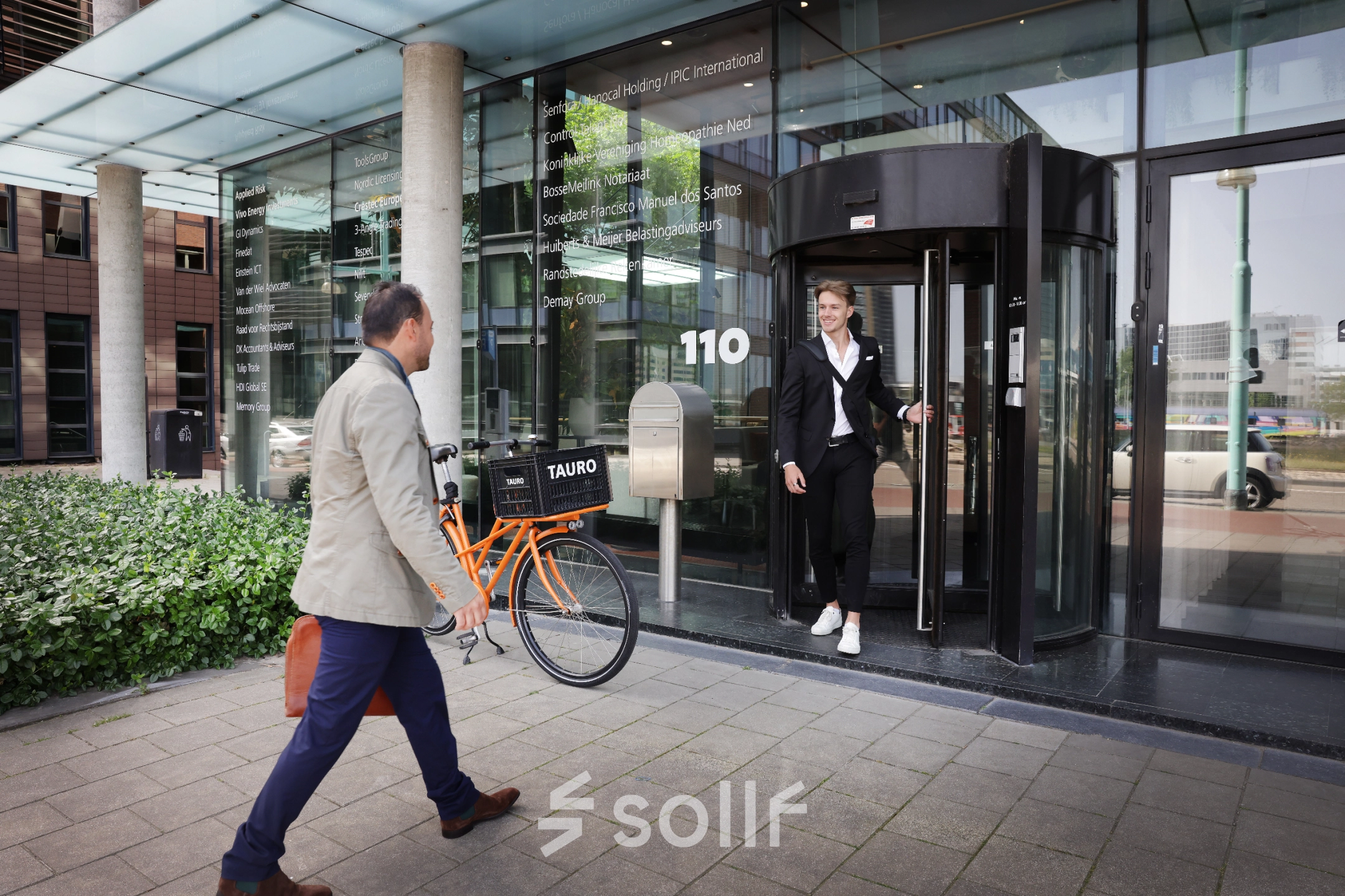 Twee personen voor het kantoorpand op Teleportboulevard 110 in Amsterdam Sloterdijk, een persoon komt naar buiten terwijl een ander met een fiets richting de ingang loopt.