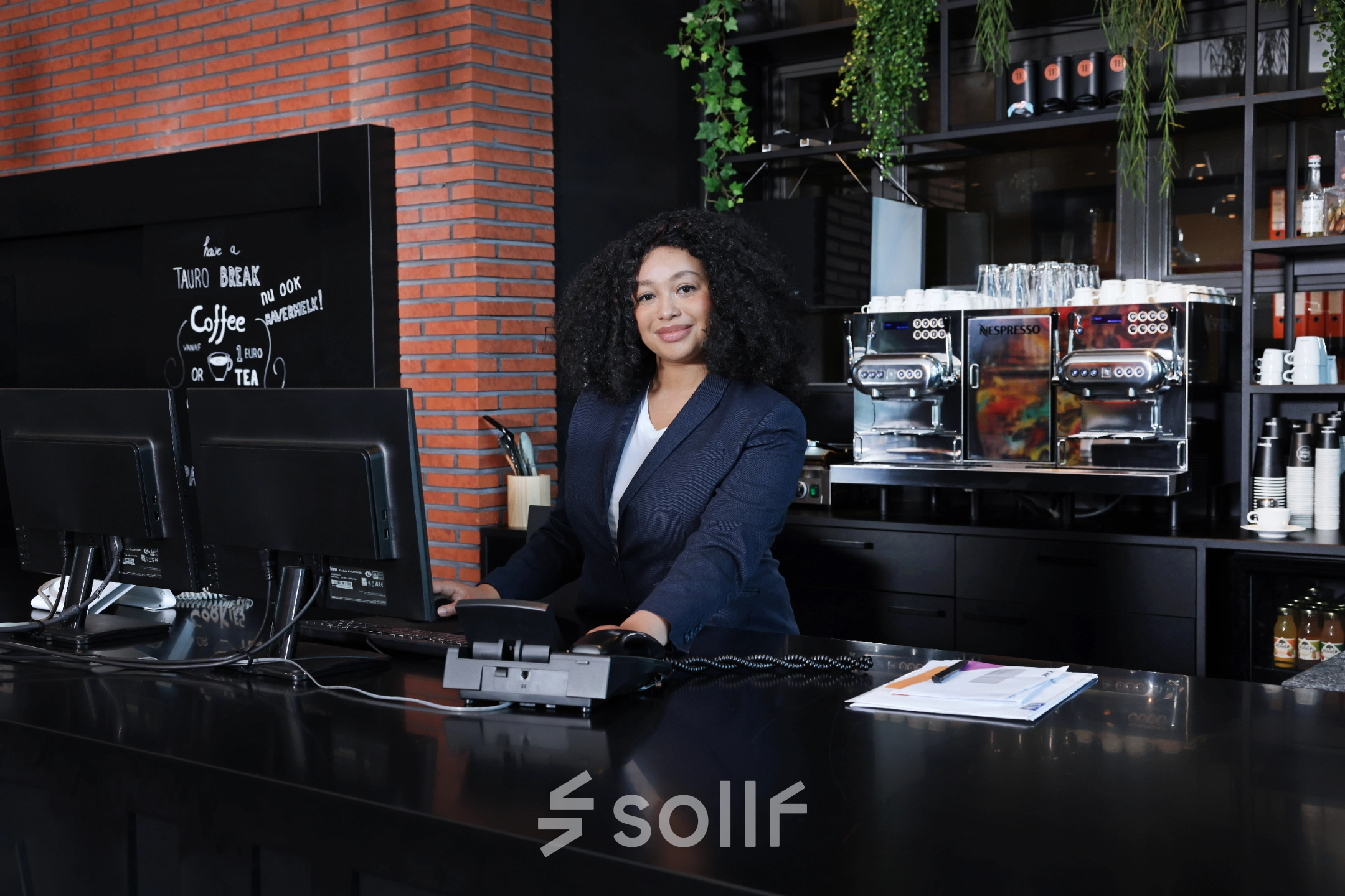 Zakelijke vrouw werkt achter de balie in een stijlvol kantoorpand op Teleportboulevard 110 in Amsterdam Sloterdijk. Op de achtergrond zijn koffiemachines en groene planten zichtbaar.