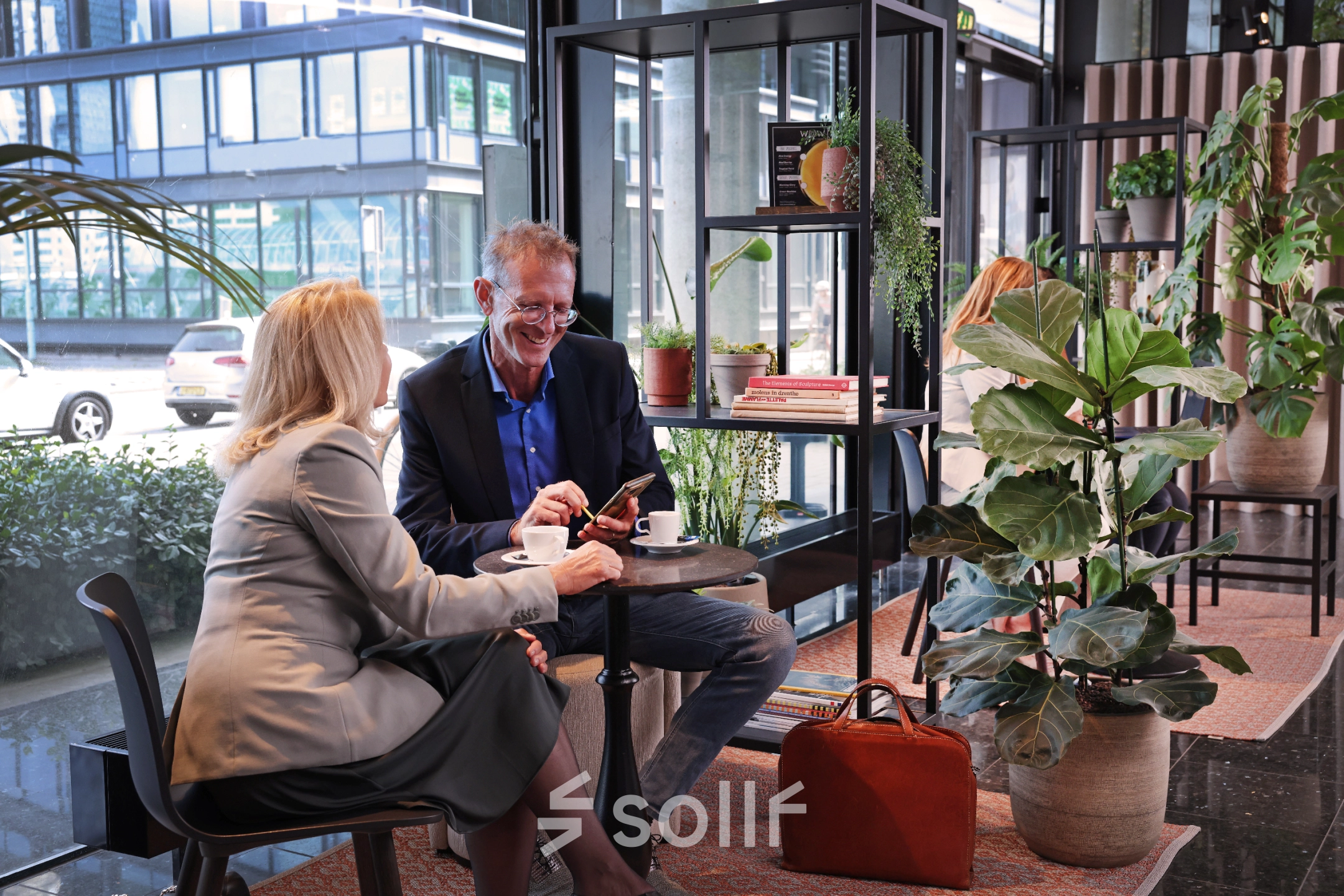 Zakelijke ontmoeting in gemeubileerde kantoorruimte op Teleportboulevard 110, Amsterdam Sloterdijk. Twee mensen overleggen met koffie, omringd door planten en moderne meubels.