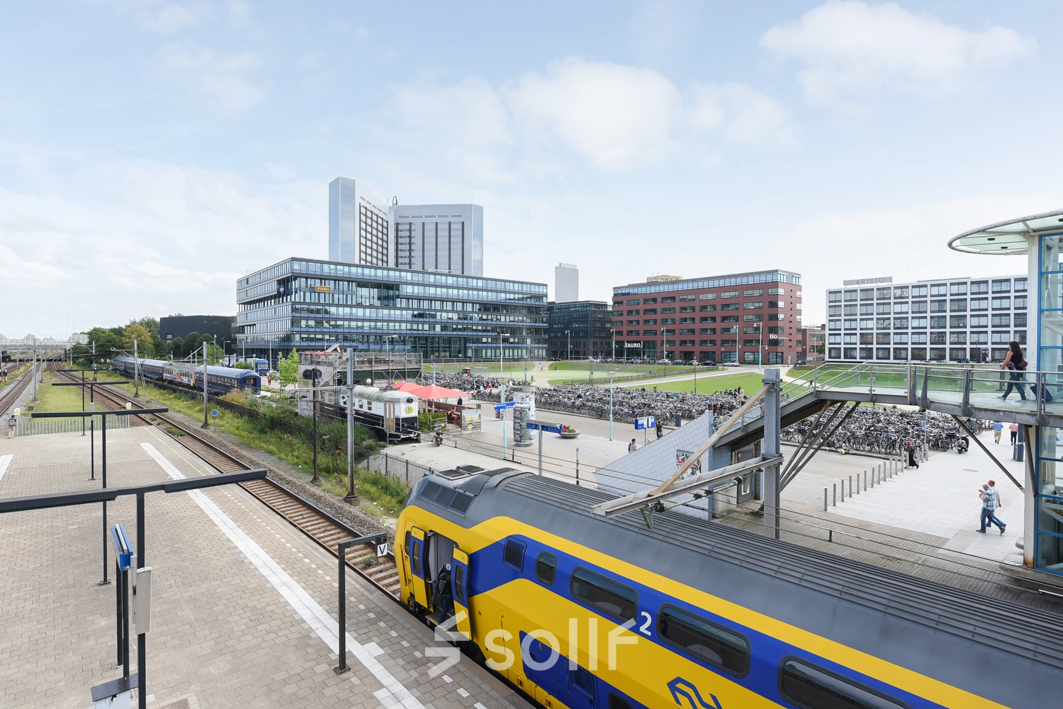 Buitenzijde van kantoorpand op Teleportboulevard 110 in Amsterdam Sloterdijk, met nabijgelegen treinsporen en een gele NS-trein. Ideale locatie voor kantoorruimte huren met uitstekende openbaar vervoer verbindingen.