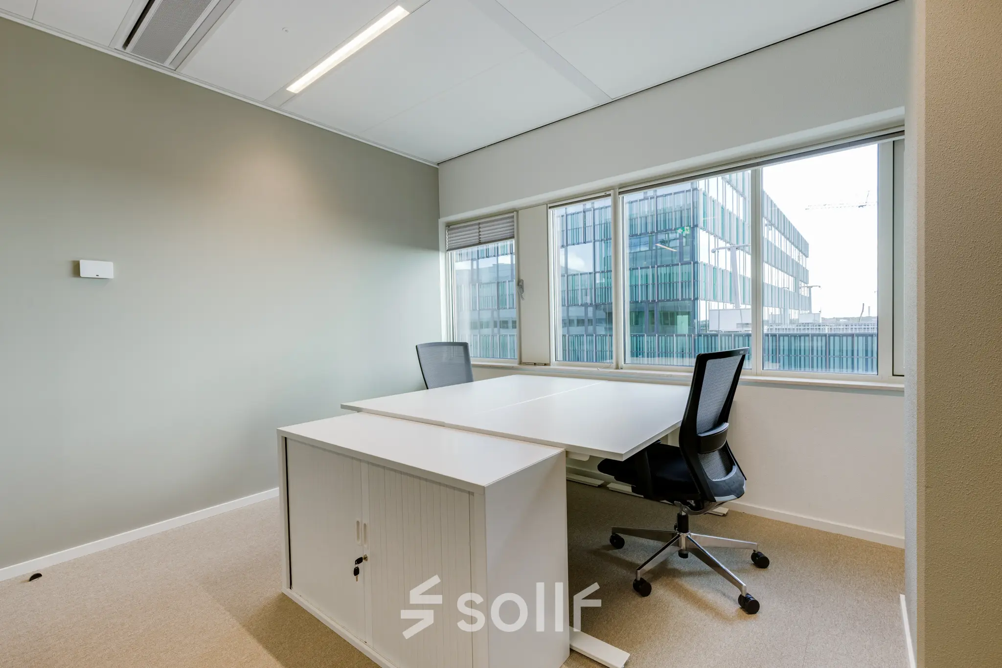 Kantoorruimte met twee bureaus en stoelen bij een raam in een kantoorpand op Herikerbergweg 288, Amsterdam Zuid-Oost. Ideaal om te huren als werkplek met veel daglicht.