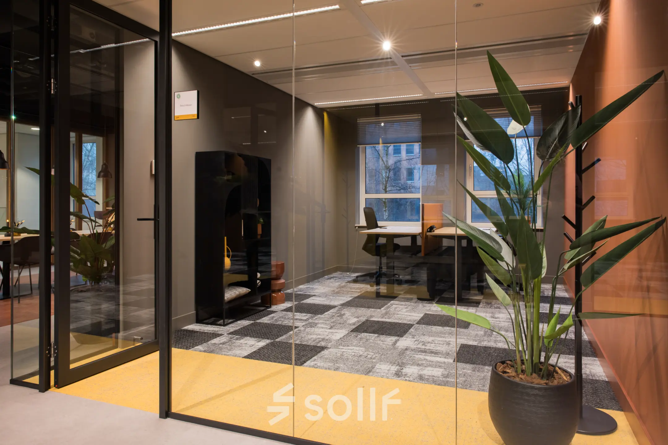Moderne kantoorruimte huren in Amsterdam Zuid-Oost met glazen wanden en grijze vloertegels. Voorzien van een bureau, stoel en grote kamerplant, geschikt voor professionele activiteiten.
