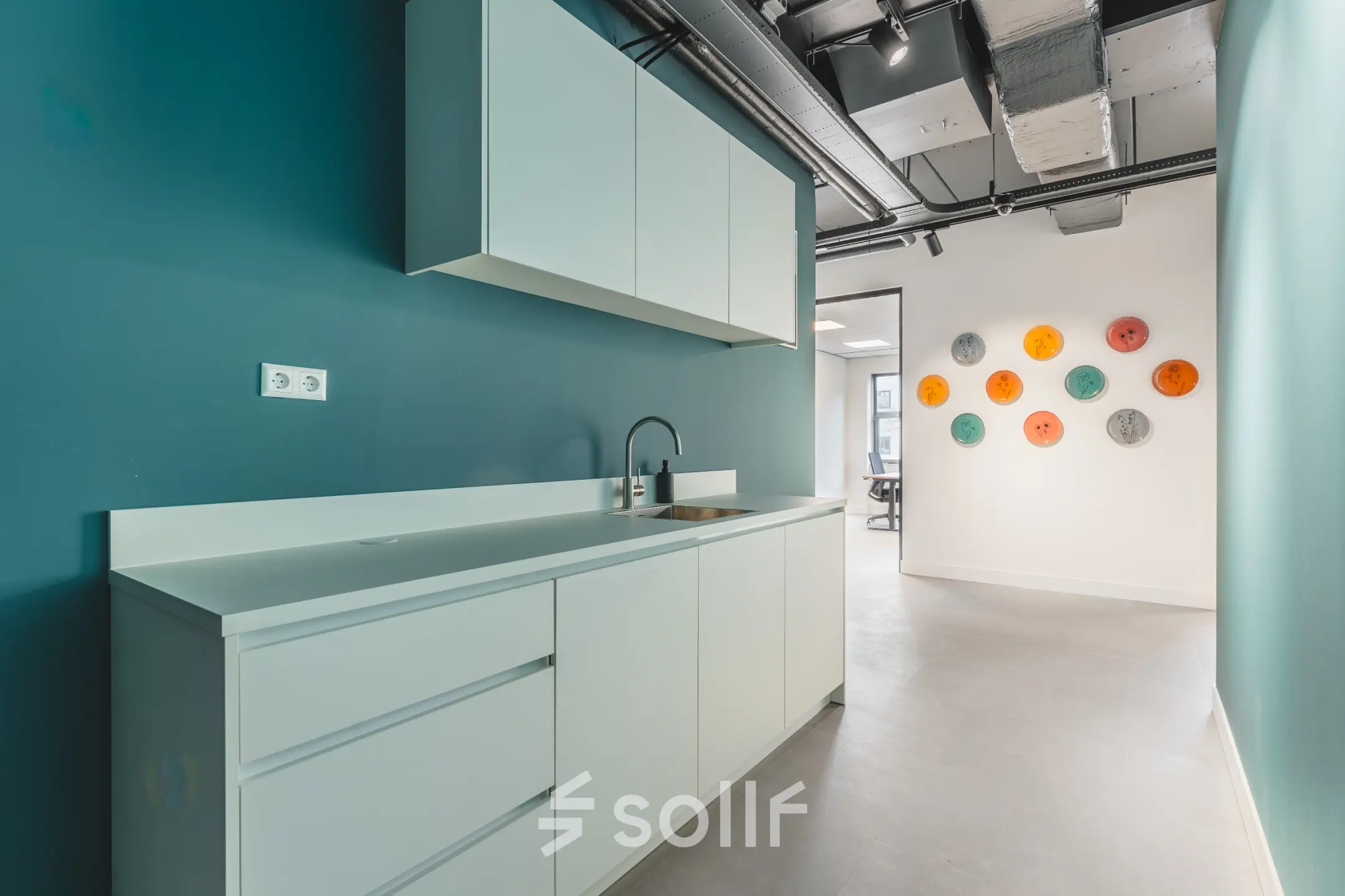 Moderne pantry in het kantoorpand op Paasheuvelweg 20, Amsterdam Zuid-Oost, met witte kasten en een blauwgroene muur. Voorzijde met wasbak en wanddecoratie in diverse kleuren. Ideaal voor kantoorruimte huren.