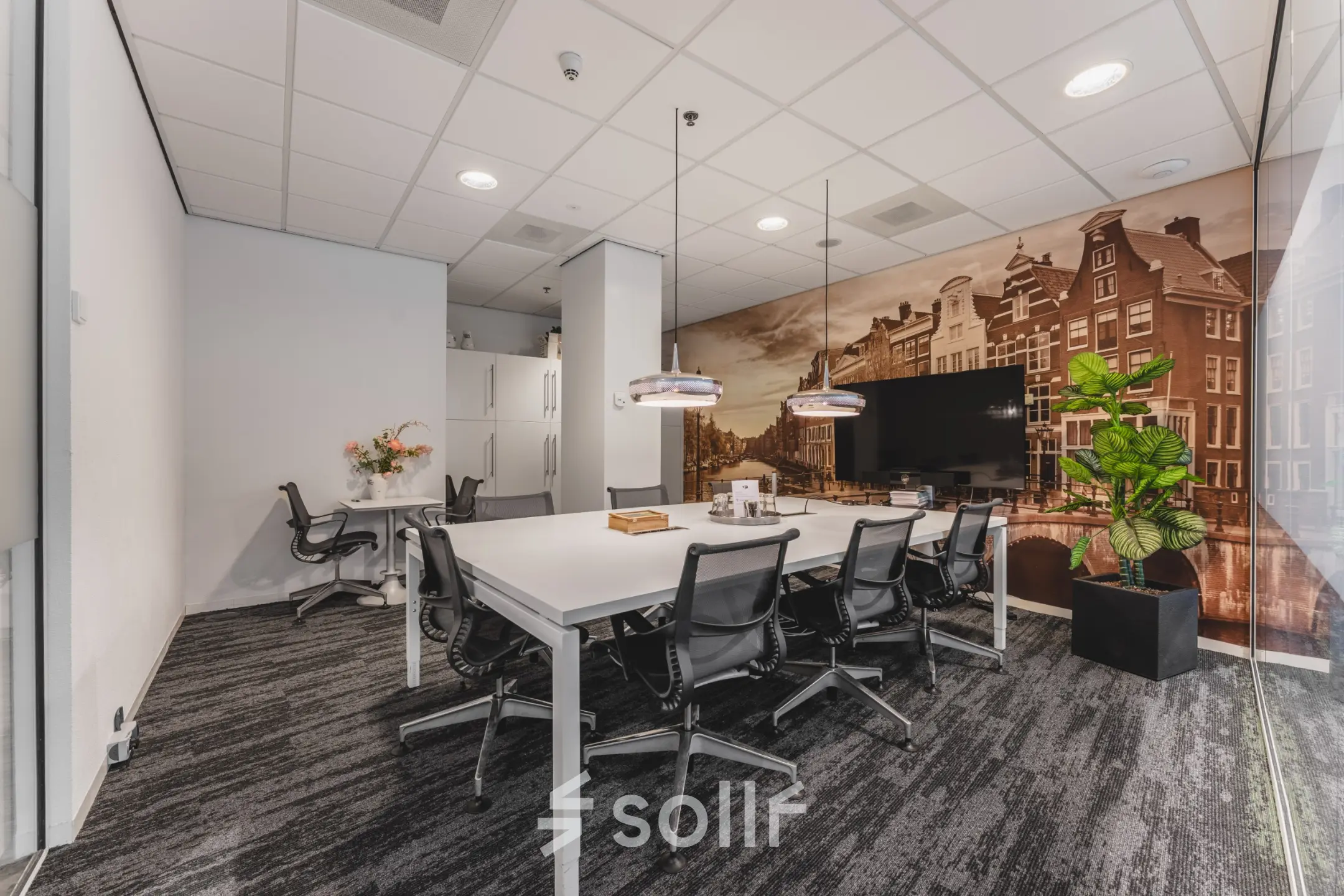 Moderne kantoorruimte te huur op Claude Debussylaan 10, Amsterdam Zuidas, met grote vergadertafel en stijlvolle stoelen, een decoratieve muur en een plant.