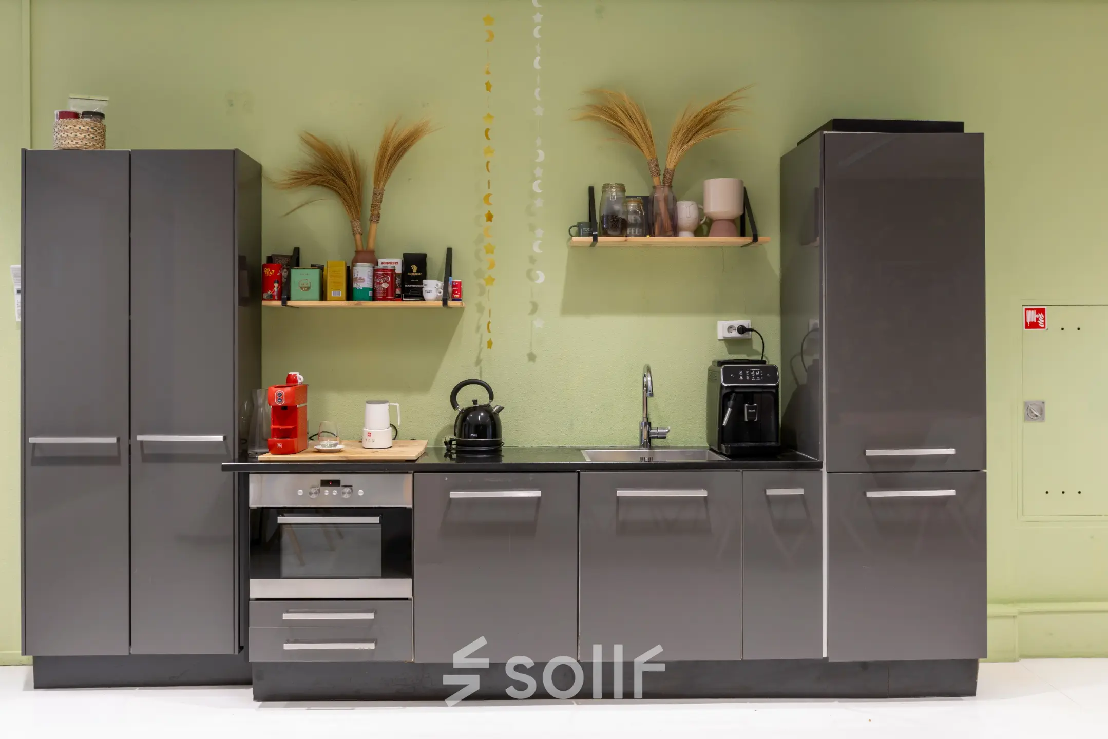 Gemeubileerde pantry van een kantoorpand met moderne grijze kastjes, een koffiezetapparaat en decoratieve elementen. Ideaal voor wie kantoorruimte wilt huren in Baarn.