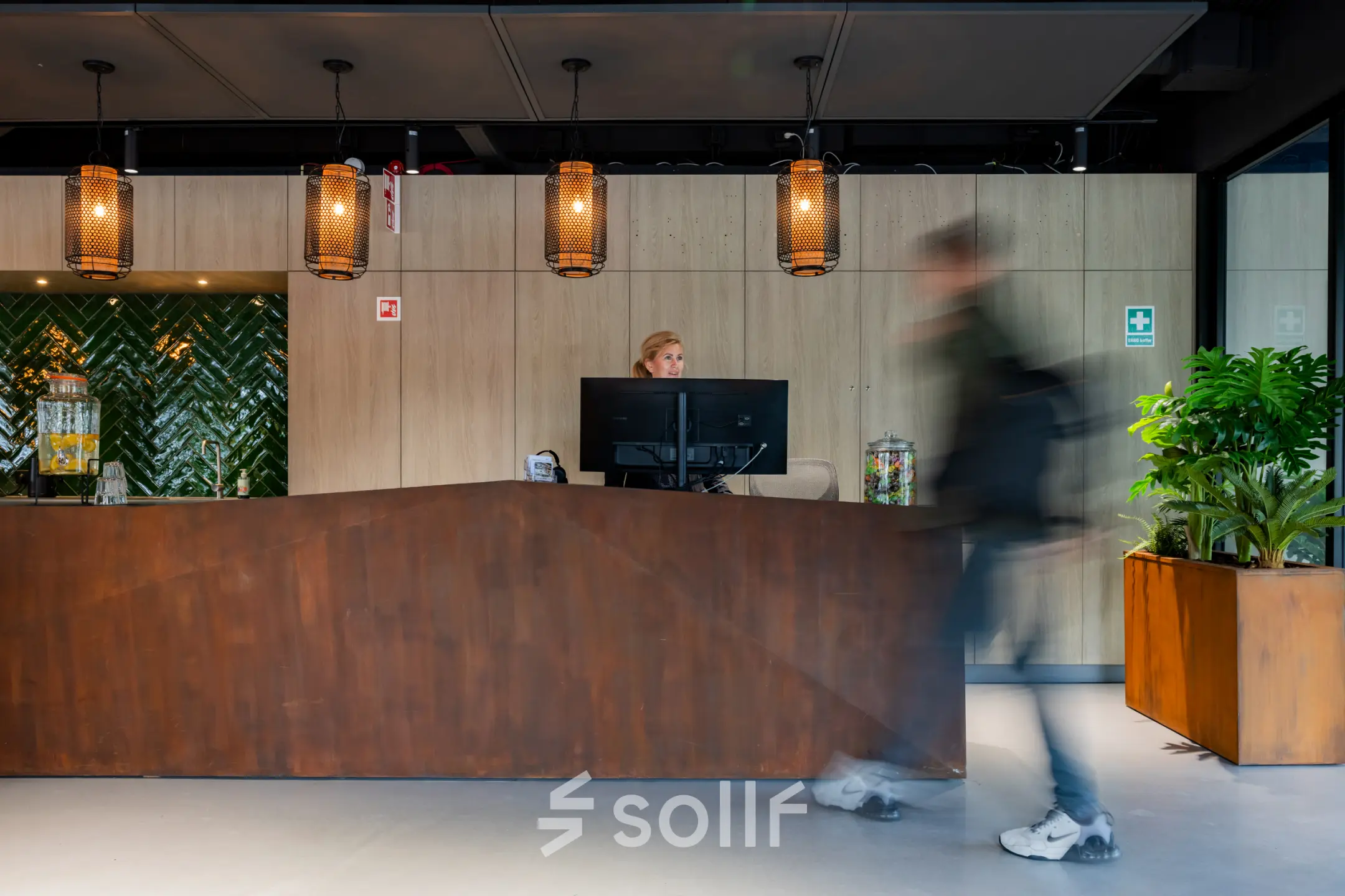 Moderne receptie in kantoorpand aan de Baron van Nagellstraat 136, Barneveld met houten balie, groen tegelwerk en planten. Persoon staat achter de balie en verwelkomt bezoekers in de kantoorruimte.