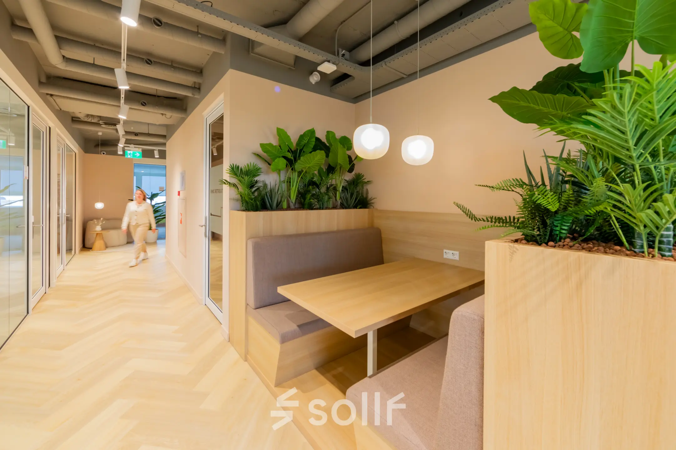 Moderne kantoorruimte huren in Barneveld op Baron van Nagellstraat 136 met groene planten, houten tafels en vloeren. Een persoon loopt door de gang.