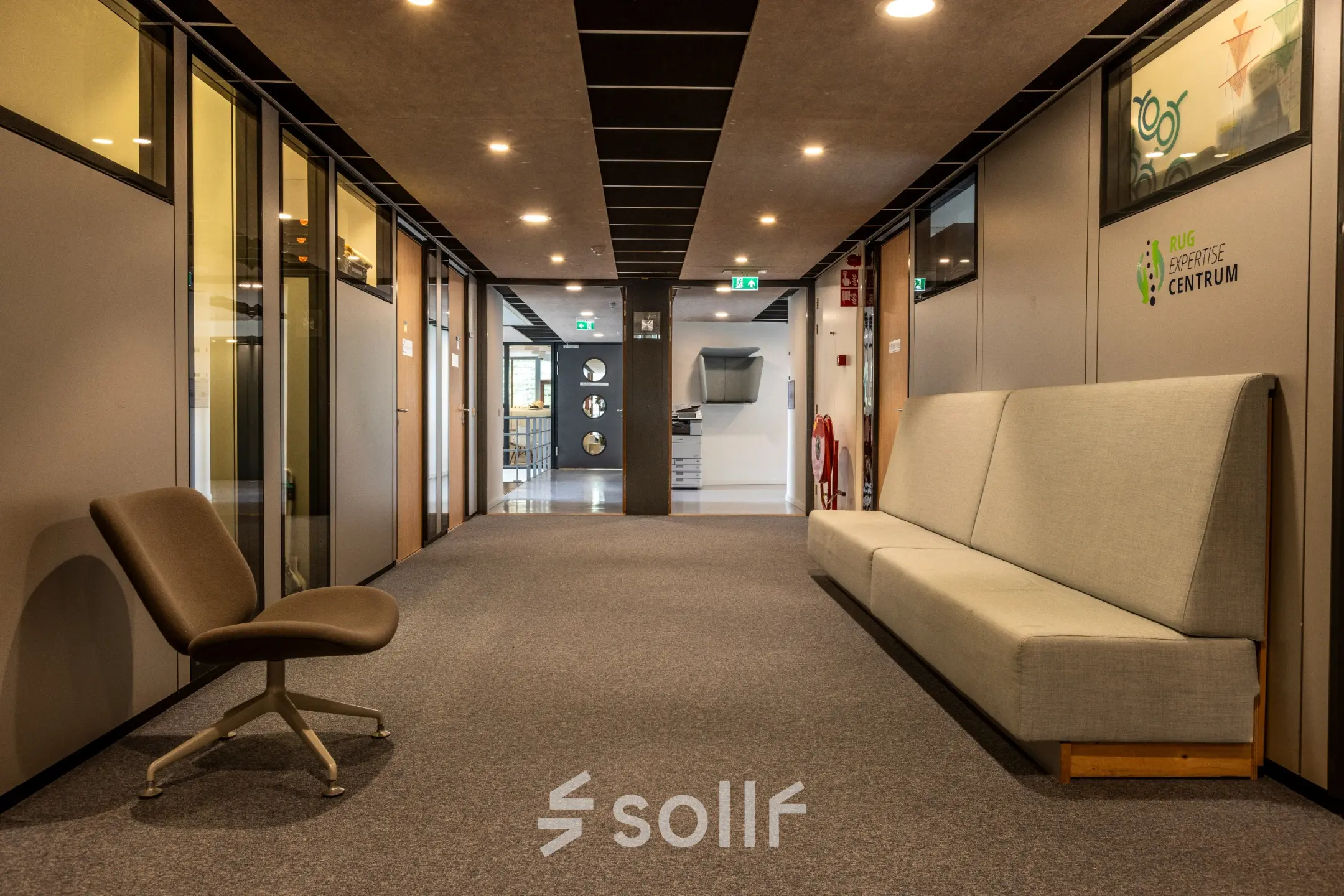 Kantoorruimte huren op Delftechpark 9 in Delft, met moderne grijze stoelen en banken in een ruime gang. Verlichte plafonds geven een heldere uitstraling aan de kantoorgang.