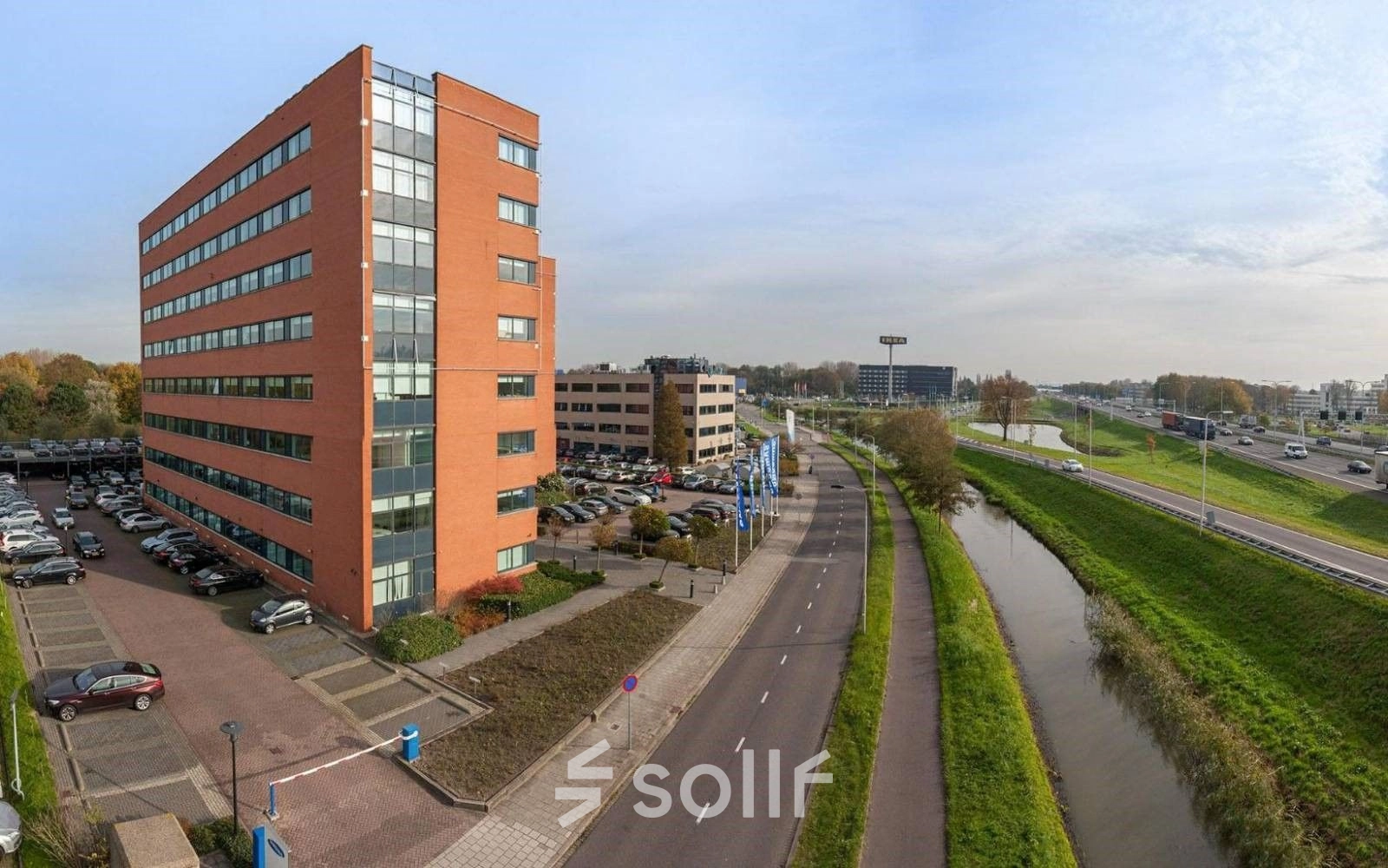 Kantoorpand aan de Olof Palmestraat 10 in Delft, met meerdere parkeerplaatsen voor het gebouw langs een rustige weg en een aangrenzende sloot.