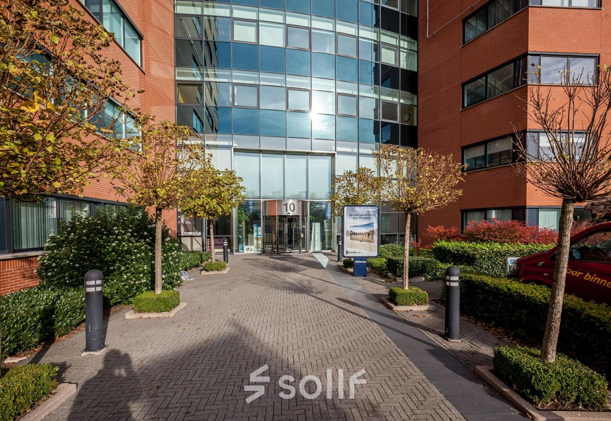 Toegang tot kantoorgebouw aan de Olof Palmestraat 10 in Delft, met moderne glazen entree omgeven door bakstenen gevel en groenvoorzieningen. Ideale locatie voor kantoorruimte huren.