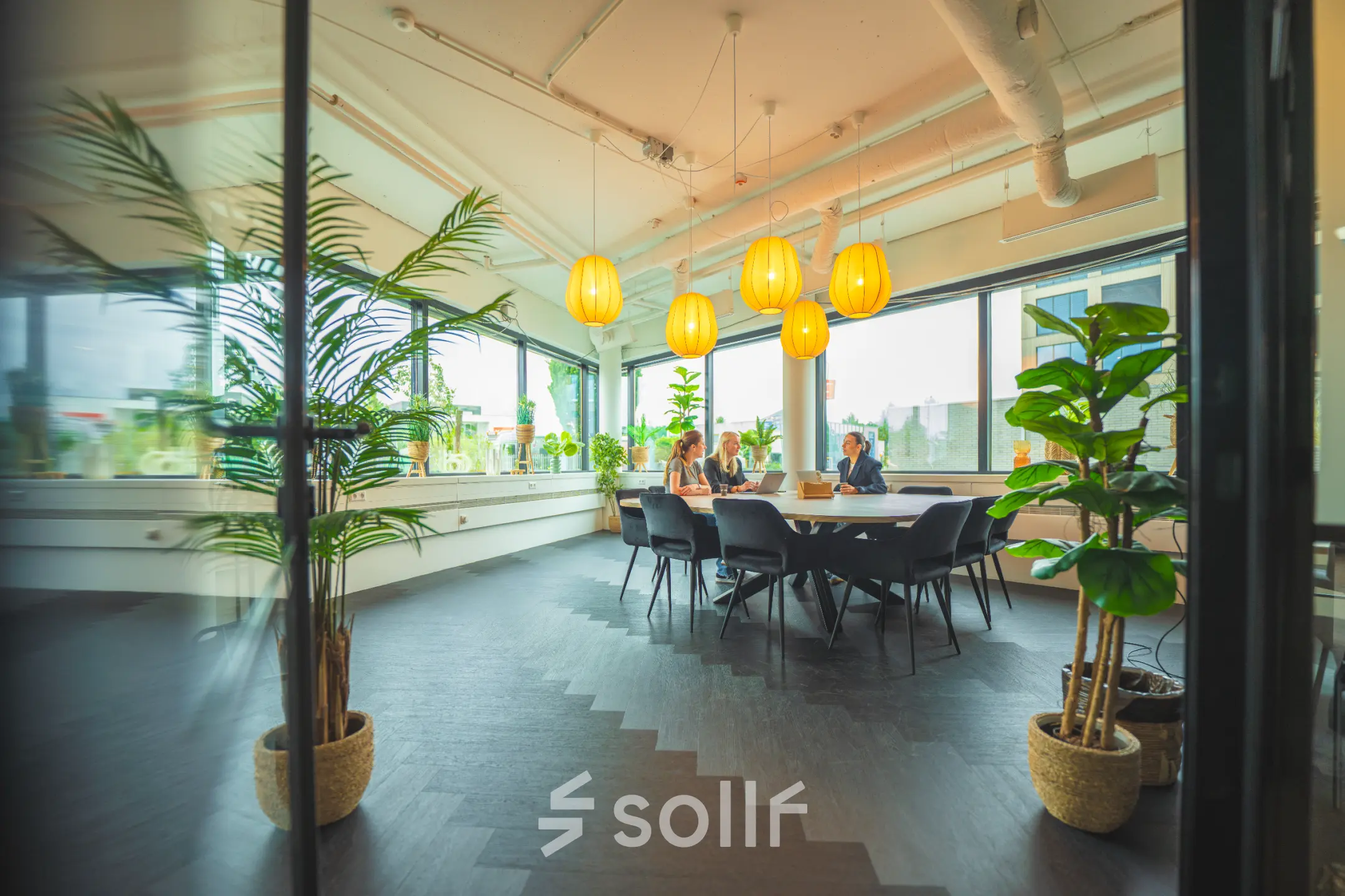 Lichte kantoorruimte met grote ramen, zes hanglampen en tafels met stoelen, gelegen op Hambakenwetering 1, Den Bosch. Twee personen zijn bezig met een zakelijke bespreking.