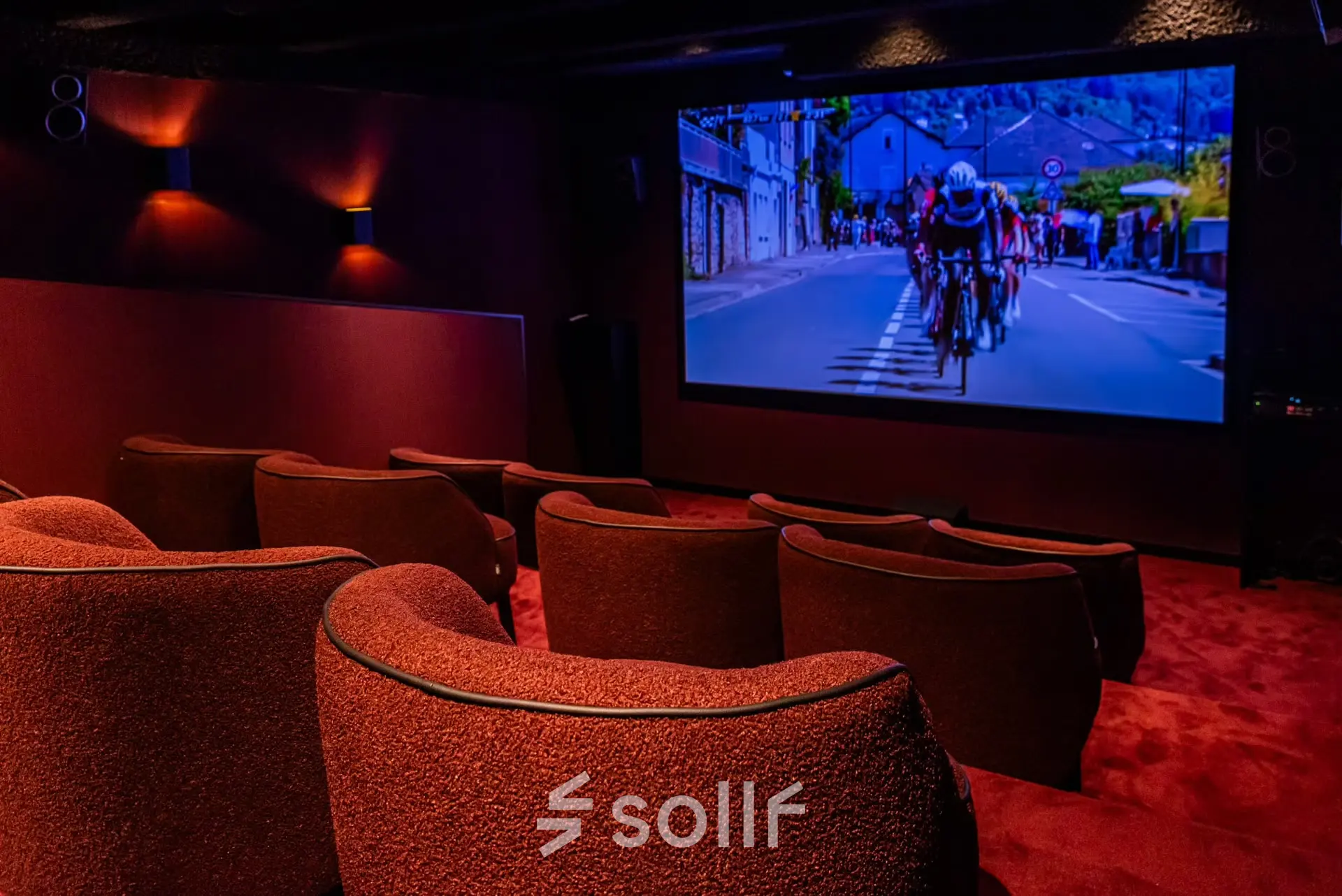 Een stijlvolle, donkere bioscoopzaal met comfortabele rode stoelen en een groot scherm met een wielerwedstrijd, ideaal voor ontspanning of presentaties in het kantoorpand op Reitschweg 1 in Den Bosch.